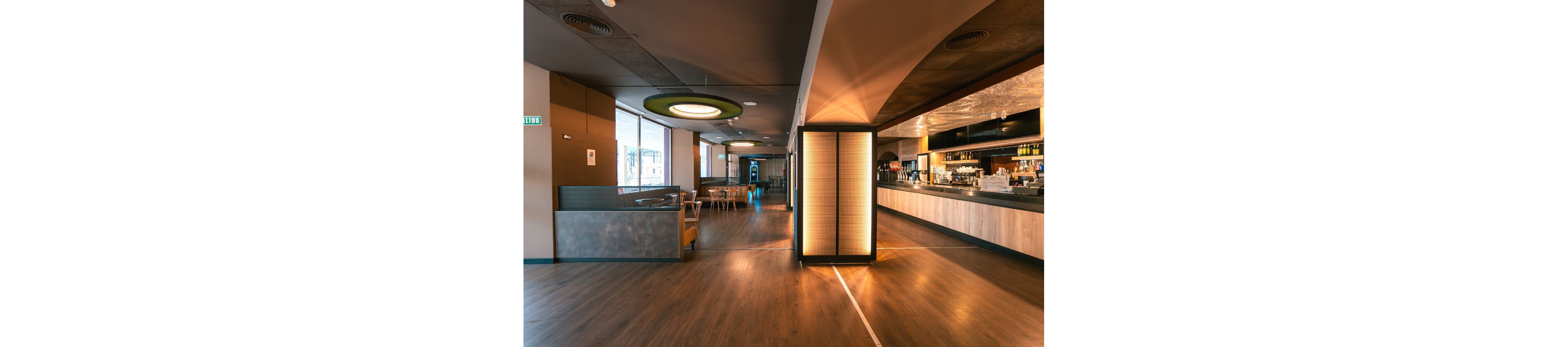 A contemporary bar lounge interior features a long counter and neatly arranged seating areas. Warm ceiling lights and a glowing central column create a cozy atmosphere across the wooden floor. Large windows on the left bring in natural light, while the bar shelves and equipment are visible in the background. The space appears clean, organized, and ready to welcome guests.
