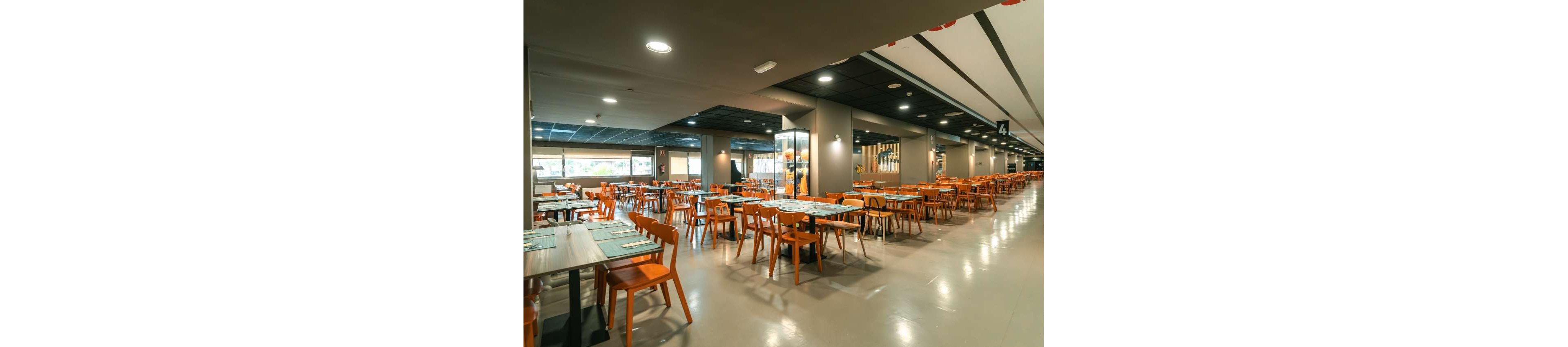 A spacious modern cafeteria is shown with rows of wooden tables and orange chairs neatly arranged. The room is brightly lit with a combination of natural light from large windows and ceiling fixtures. Place settings and placemats are prepared on the tables, but no people are present. Neutral walls, polished floors, and a long corridor create a clean, organized dining environment.