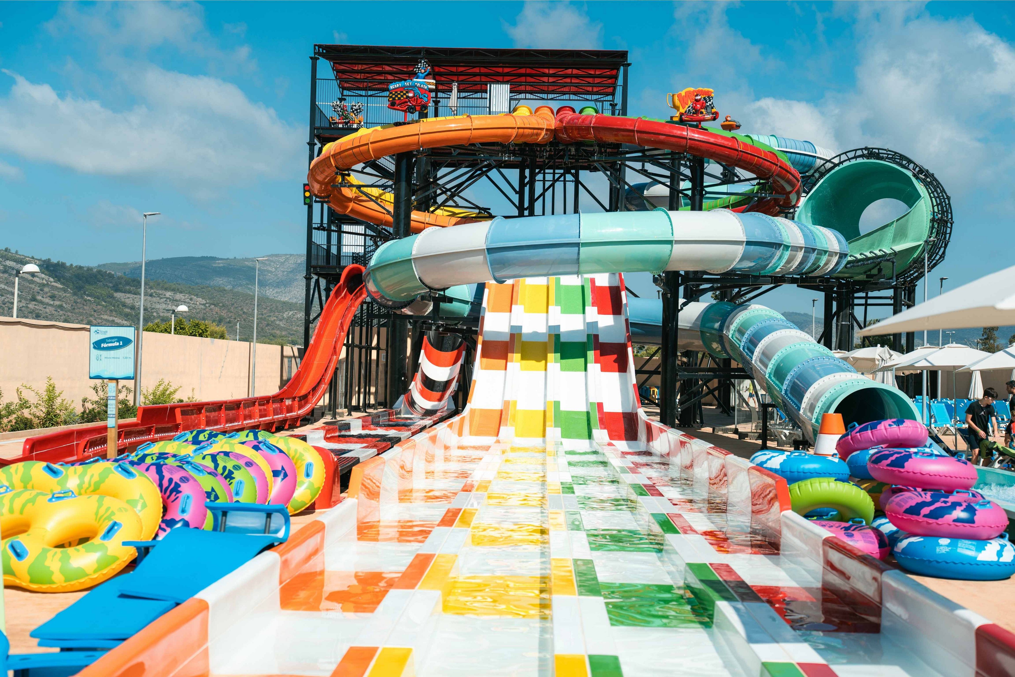 A large, colorful multi-lane water slide dominates this outdoor water park scene. Bright inflatable tubes are stacked near the base of the slide, ready for visitors. The structure includes twisting enclosed tubes and open slides in various colors, set against a backdrop of hills and blue sky. Sunshades and seating areas are visible to the side, suggesting a family-friendly recreational environment.