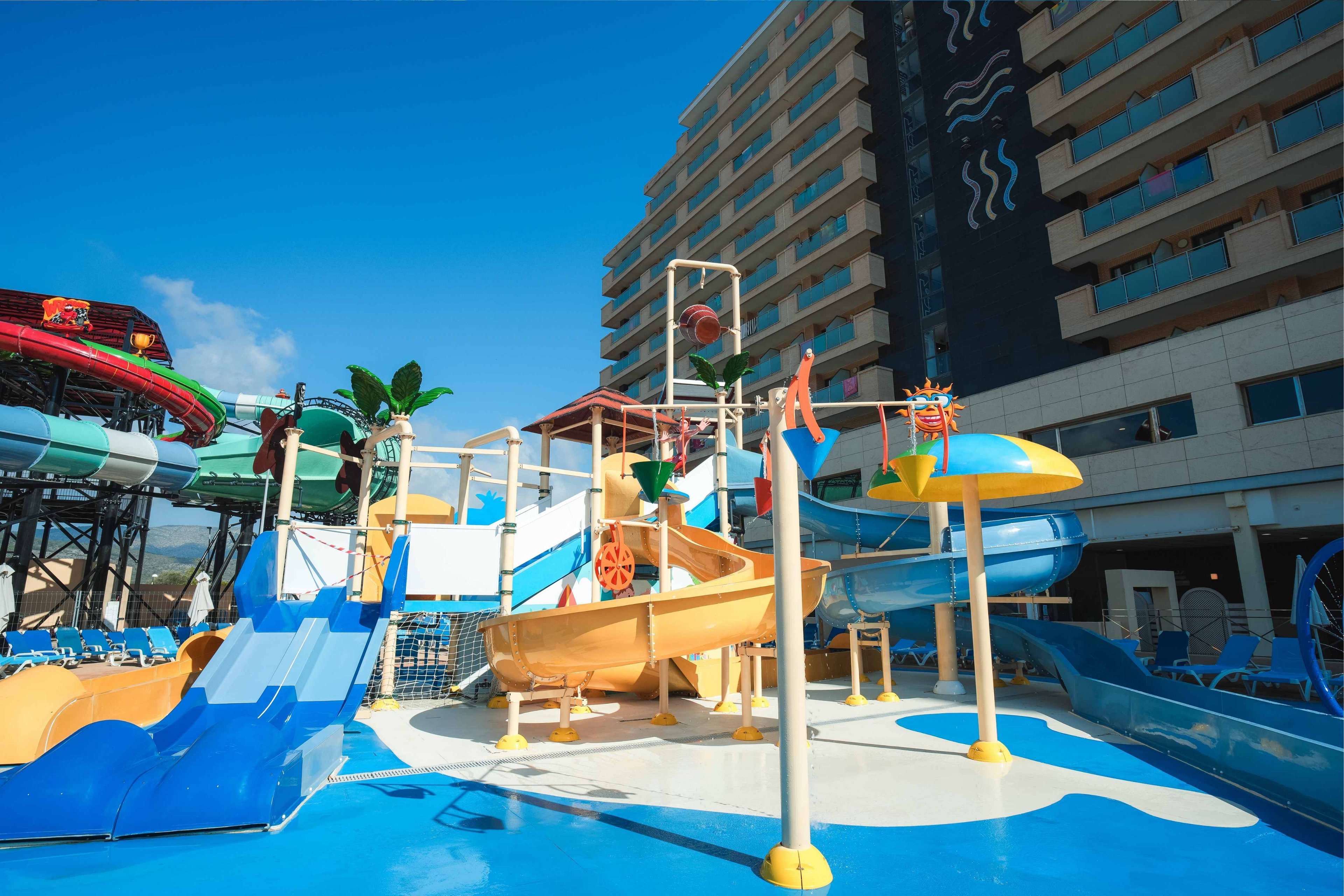 A bright children’s water play area features multiple slides and splash structures under a clear blue sky. The scene includes a shallow pool, small water slides, and playful elements like palm tree decorations and a tipping bucket. A large hotel or resort building rises in the background, suggesting an urban or seaside vacation setting. The vivid colors and sunny weather create a fun, family-friendly atmosphere.