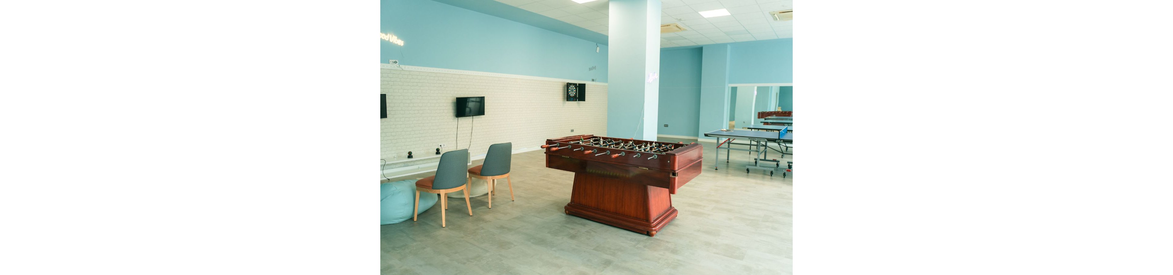 A spacious recreation room features a large wooden foosball table as the central focus. The setting includes light blue walls, a tiled floor, and ample open space for movement. In the background, there are chairs, wall-mounted screens, a dartboard, and a ping pong table, suggesting multiple entertainment options. The overall atmosphere is bright, casual, and suitable for socializing or relaxing.