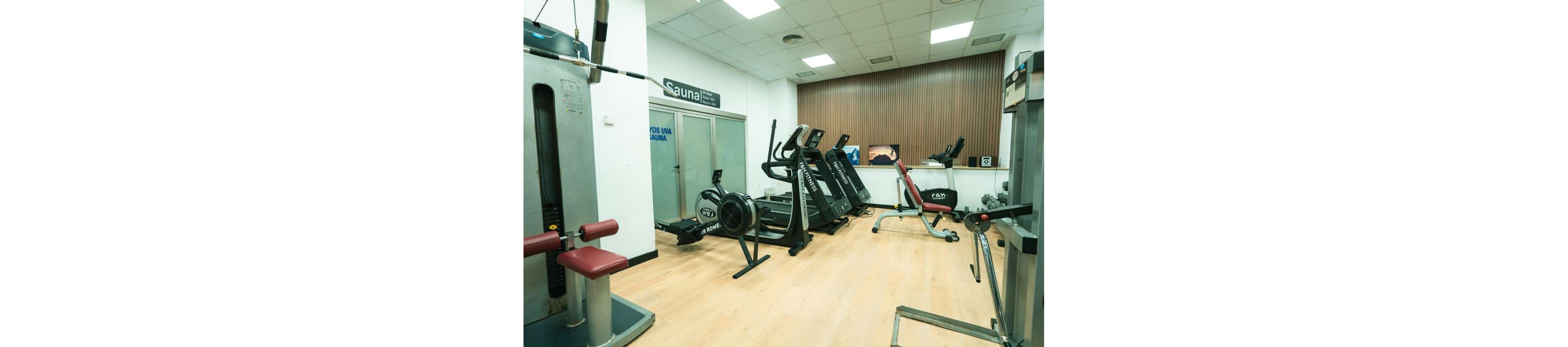 A clean, modern gym room features several cardio machines including treadmills, an exercise bike, and a rowing machine arranged along a wall. Strength training equipment such as a weight machine and an adjustable bench are also visible on the light wooden floor. Glass doors in the background display a sign reading "Sauna" and another sign with the text "FOG UMA SAUNA." The space is brightly lit with ceiling lights and has a minimalist, functional design.