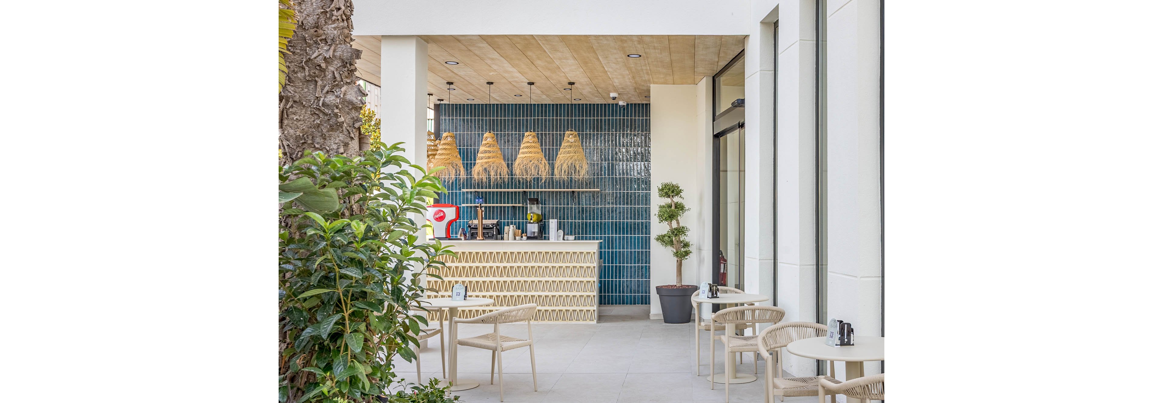 A contemporary outdoor café features a small coffee counter set against a blue tiled wall. Woven pendant lights hang above the bar, adding warmth to the clean, modern design. Light-colored chairs and round tables are arranged on a spacious patio beside large glass windows. Green plants and trees soften the architecture, creating a relaxed and inviting atmosphere.
