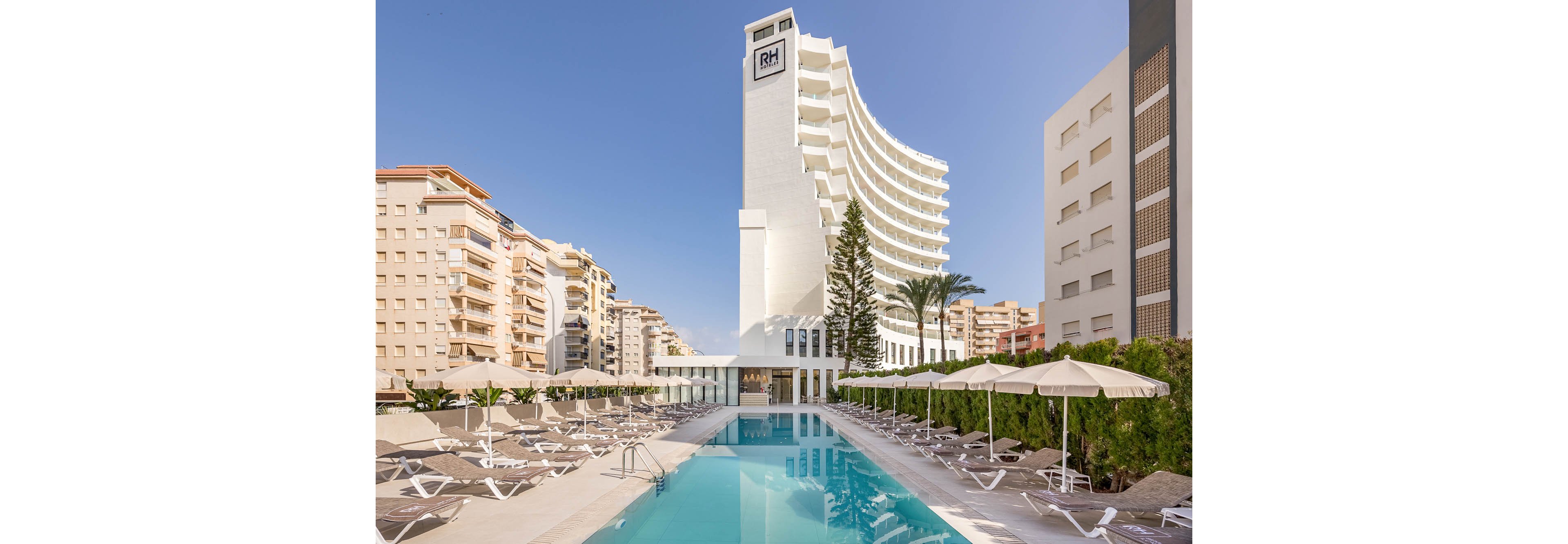 A tall white hotel tower rises above a long rectangular outdoor swimming pool in an urban coastal setting. The pool is lined with empty sun loungers and white umbrellas, suggesting a resort environment. Surrounding apartment buildings frame the scene under a clear blue sky. The hotel facade is minimalist and modern, with a prominent RH sign near the top of the tower.