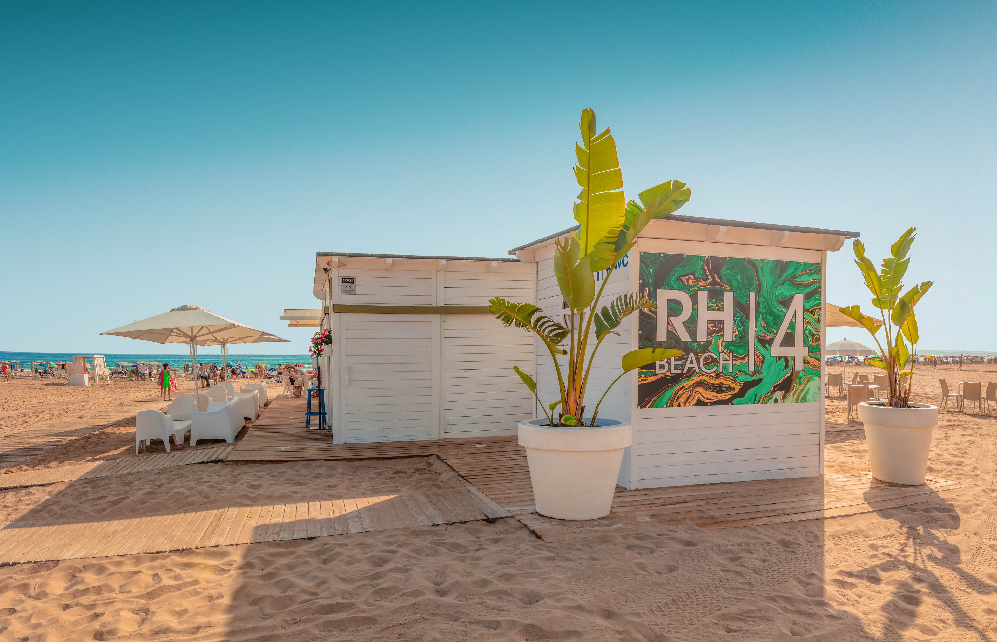 A white beach bar hut stands on a sandy shoreline under a clear blue sky. Large potted tropical plants frame a colorful sign that reads RH4 BEACH with a leafy pattern. Sun loungers and umbrellas are visible in the background near the sea, suggesting a relaxed seaside atmosphere. The warm lighting and soft shadows evoke a calm, summery mood.