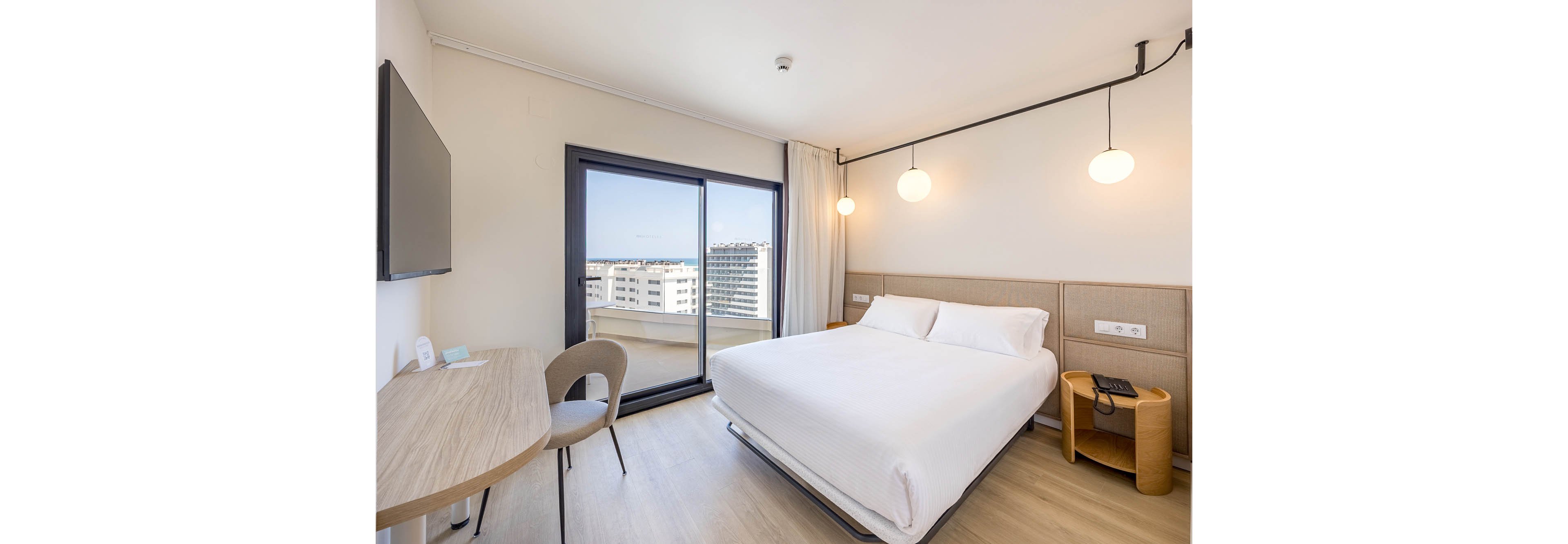 A bright modern hotel room features a neatly made double bed with a padded headboard. To the left, a minimalist desk and chair face a wall-mounted flat-screen television. Large sliding glass doors open to a balcony overlooking several mid-rise apartment buildings and a distant skyline. Warm pendant lights and light wood flooring create a clean, contemporary atmosphere.