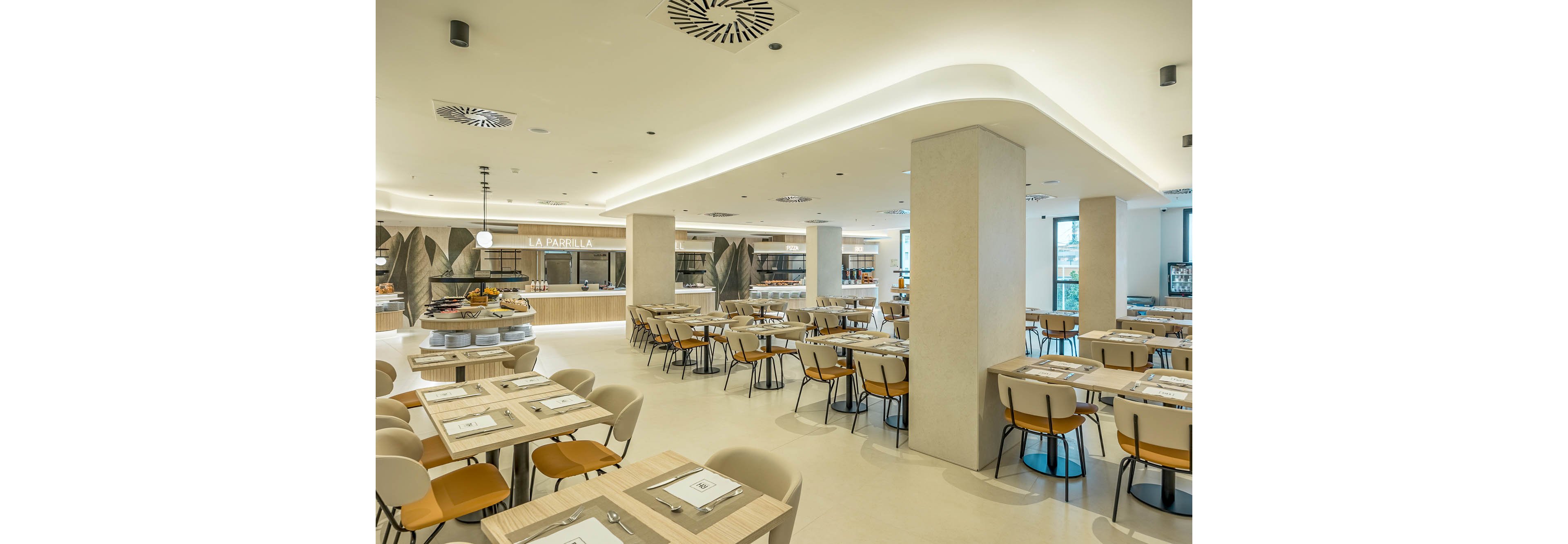 A spacious modern restaurant dining area features neatly arranged wooden tables and upholstered chairs. The setting appears to be a hotel or buffet-style restaurant with a central food station and open serving counters in the background. Neutral tones, soft lighting, and clean architectural lines create a bright and contemporary atmosphere. Large columns and ceiling lighting elements add structure and visual interest to the open-plan space.