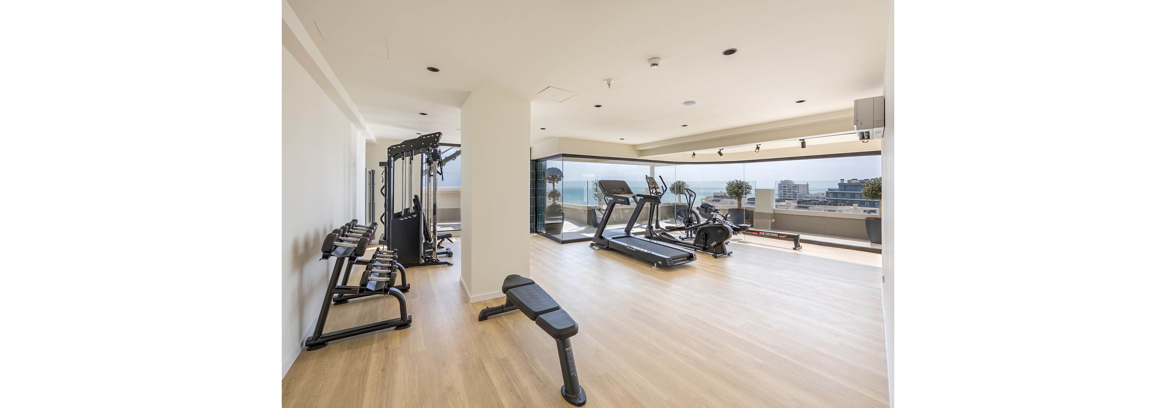 A bright, modern gym features various exercise machines arranged on a light wooden floor. Treadmills, a weight bench, dumbbells, and a multi-gym station are positioned facing large floor-to-ceiling windows. The space overlooks a cityscape and ocean, creating an open, airy atmosphere. Neutral colors and minimalist design emphasize cleanliness and contemporary style.