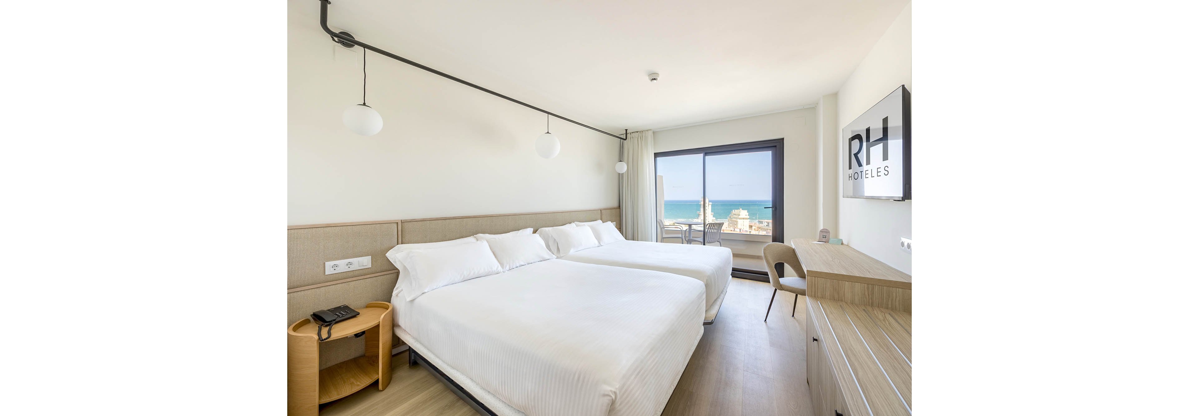 A bright, modern hotel room features two neatly made double beds with white linens. The space includes a wooden desk with a chair, a wall-mounted television displaying the text "RH HOTELES," and a bedside table with a telephone. Large sliding glass doors open to a balcony overlooking the sea and coastal buildings. Minimalist decor, light wood flooring, and neutral tones create a clean and relaxing atmosphere.