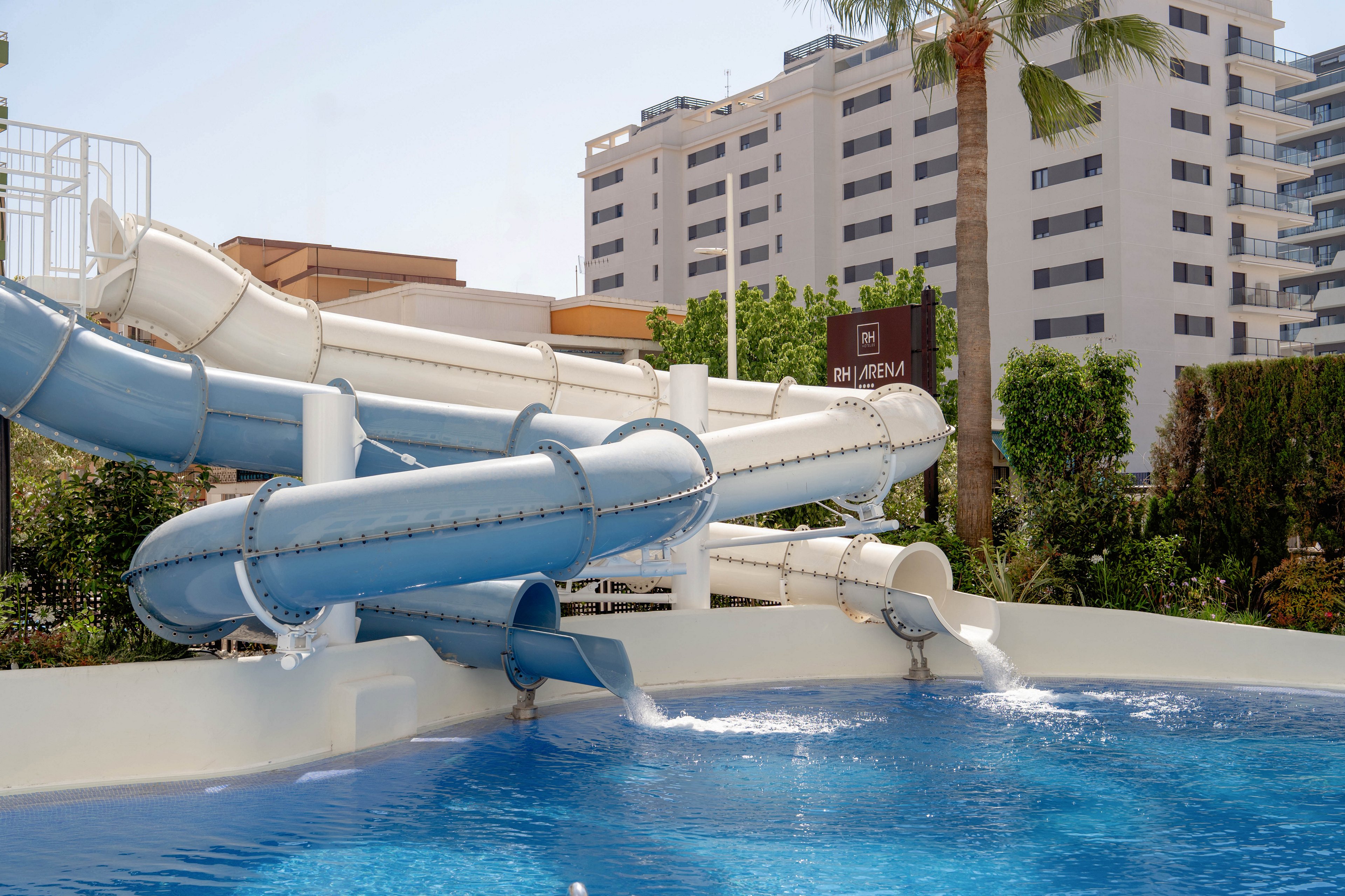 A large, twisting waterslide empties into a clear blue pool in an outdoor hotel setting. The slide features light blue and white enclosed tubes supported above the pool edge. In the background, modern multi-story buildings and lush greenery frame the scene, along with a tall palm tree. Bright daylight and calm water suggest a relaxed, off-peak moment at a resort or water park.