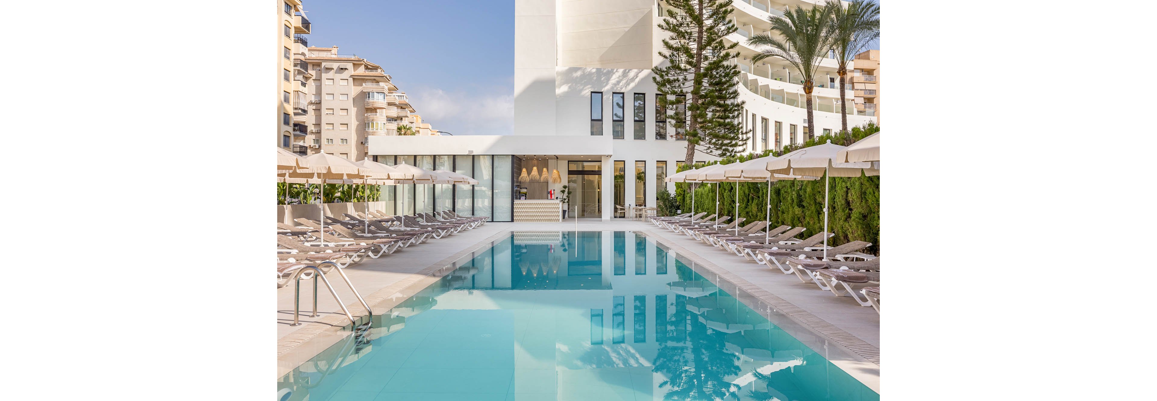 A long rectangular swimming pool stretches between rows of beige sun loungers and umbrellas at a modern hotel. The white building facade and tall windows reflect in the calm turquoise water. High-rise apartments and palm trees frame the scene, suggesting an urban resort setting. The overall atmosphere is bright, clean, and inviting, ideal for relaxation and leisure.