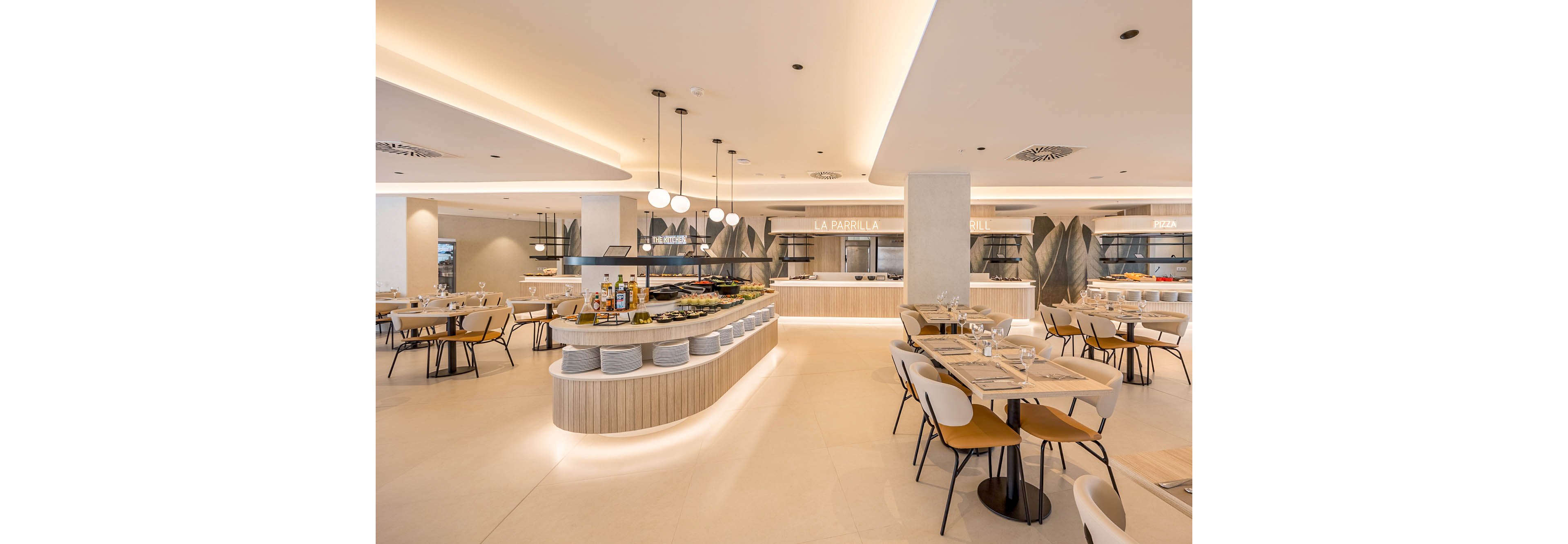 A spacious, modern buffet restaurant features a central island filled with dishes and condiments. Neatly set tables with plates, glasses, and cutlery surround the buffet area, ready for guests. Warm neutral tones, soft ceiling lighting, and clean lines create a contemporary, minimalist atmosphere. The open kitchen and service counters in the background emphasize a hotel or resort dining setting.