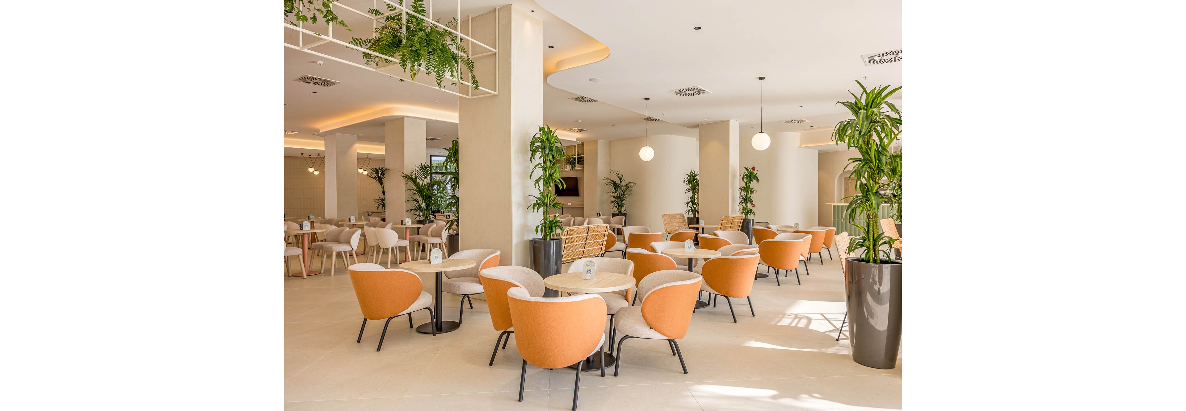 A contemporary cafe interior features round tables surrounded by stylish orange upholstered lounge chairs. The space is bright and airy, with large columns, light-colored flooring, and minimalist decor. Green potted plants and hanging greenery add a natural touch throughout the room. Warm ceiling lighting and globe pendant lights create a welcoming, relaxed atmosphere in the open seating area.