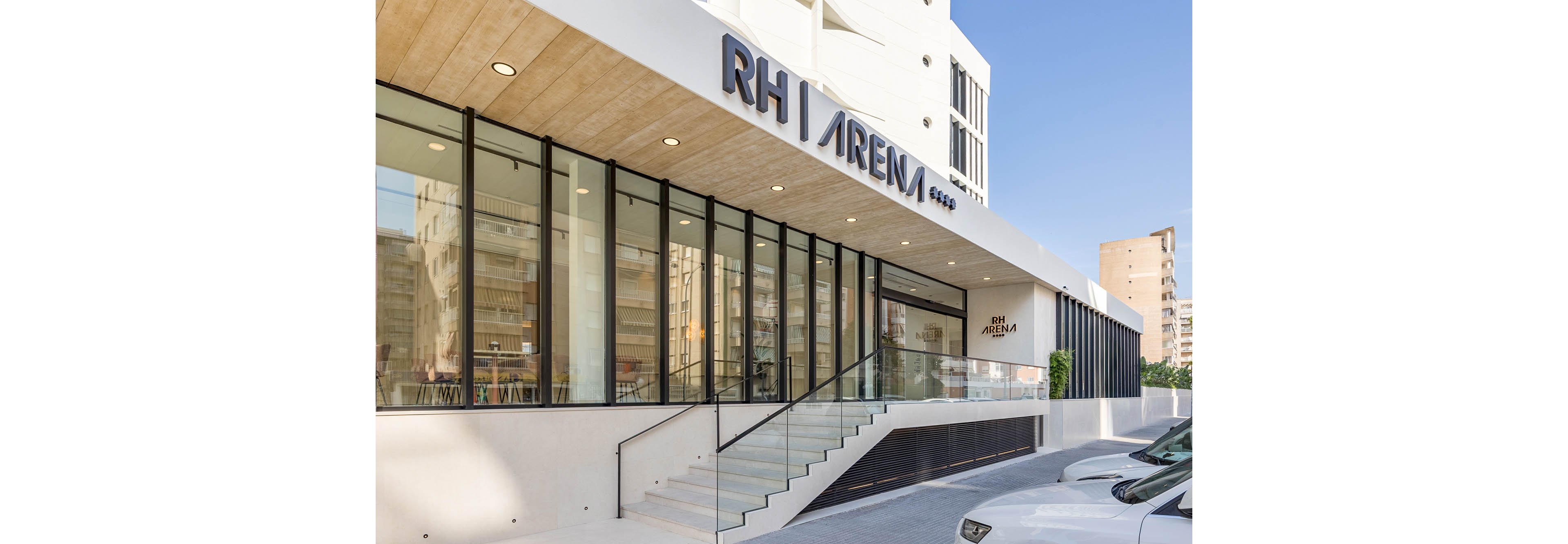 A contemporary building exterior features the clearly labeled RH Arena entrance. Large floor-to-ceiling glass windows reveal an interior with seating and a clean, minimalist design. The structure includes a light-colored overhang, a glass-railed staircase, and a smooth white facade. Urban residential buildings and parked cars are visible in the background, suggesting a city setting.