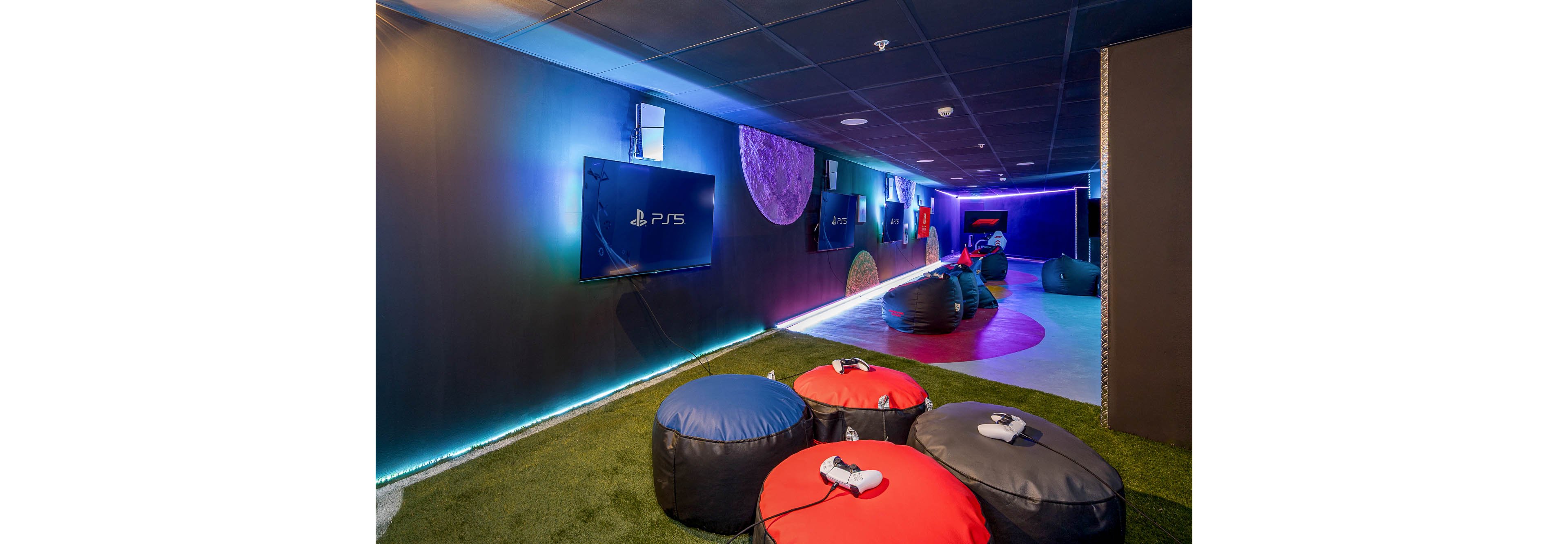 A contemporary gaming lounge features several wall-mounted PS5 screens along a long, narrow room. Colorful beanbag seats and stools are arranged on circular rugs over artificial grass flooring. Soft blue and purple LED lighting lines the walls and ceiling, creating an immersive atmosphere. Game controllers rest on the beanbags, ready for players in this relaxed entertainment space.