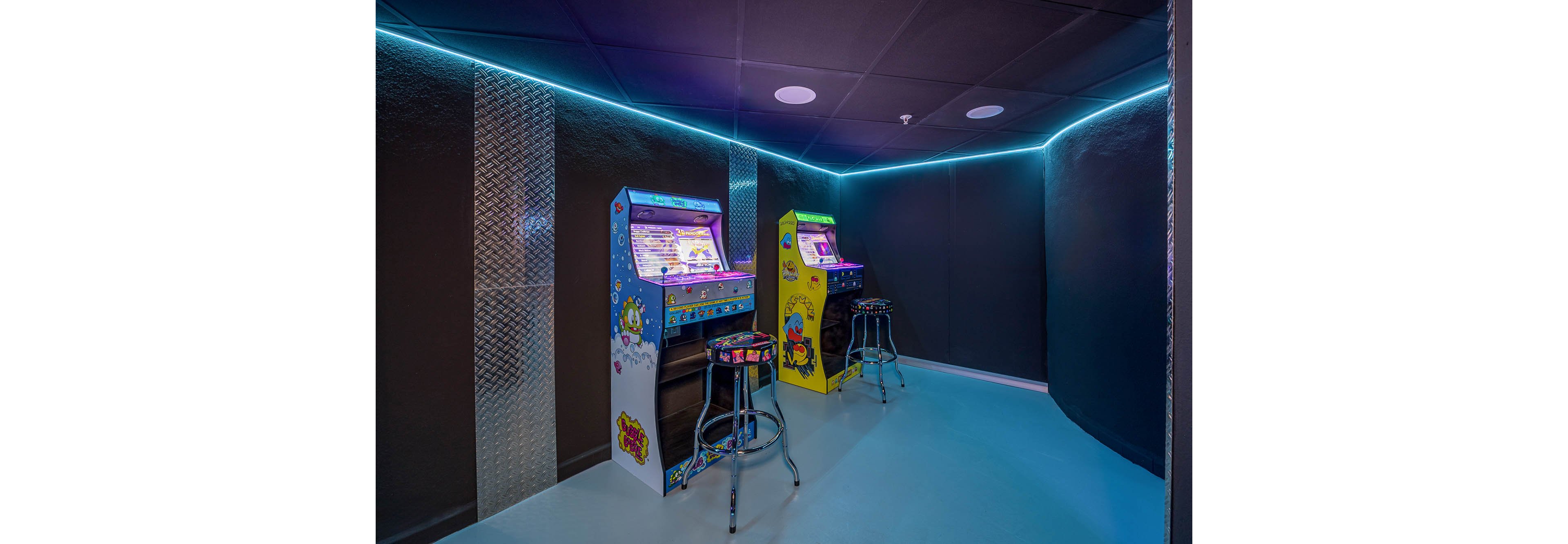 A small neon-lit room features two upright arcade game machines against a dark wall. One cabinet is blue and the other is yellow, both decorated with colorful cartoon graphics. Two chrome bar stools with patterned seats are placed in front of the machines, ready for players. The space has a modern yet nostalgic feel, with metallic wall panels and cool blue accent lighting outlining the ceiling.
