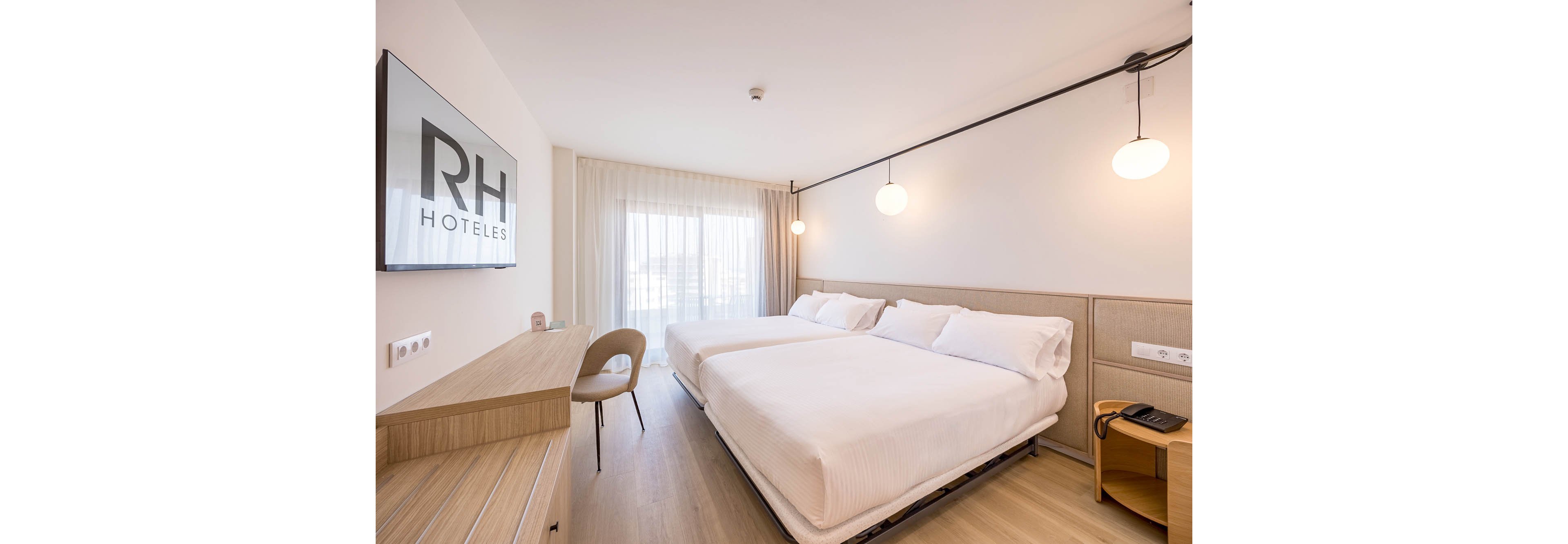A bright modern hotel room features two neatly made twin beds with white linens. The space includes a wooden desk with a chair and a wall-mounted television displaying the text RH HOTELES. Large sheer curtains cover a window or balcony door, allowing natural light to fill the room. Minimalist pendant lights and light wood flooring contribute to the clean, contemporary design. A bedside table with a telephone completes the functional guestroom setup.