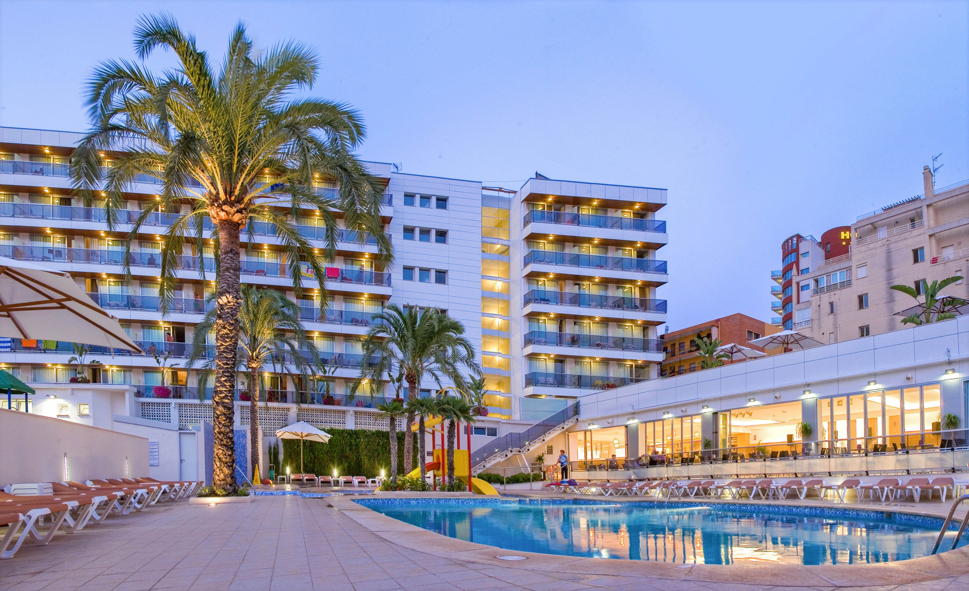A contemporary multi-story resort hotel is shown at dusk with balconies lit by warm yellow lights. In the foreground, a curved outdoor swimming pool reflects the building and the evening sky. Palm trees and neatly arranged sun loungers line the poolside terrace, creating a relaxed vacation atmosphere. Large windows on the lower level reveal a bright interior space, suggesting dining or common areas for guests.