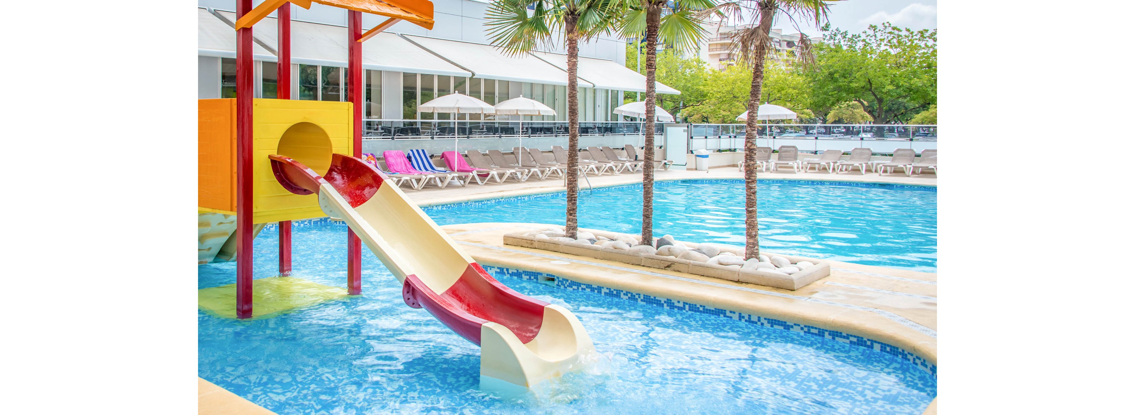 A young girl splashes into a bright blue swimming pool from a colorful waterslide. Another child waits at the top of the slide structure, ready to go next. The pool area is surrounded by lounge chairs, umbrellas, and palm trees in front of a modern hotel building. The scene suggests a sunny vacation day focused on family fun and relaxation.
