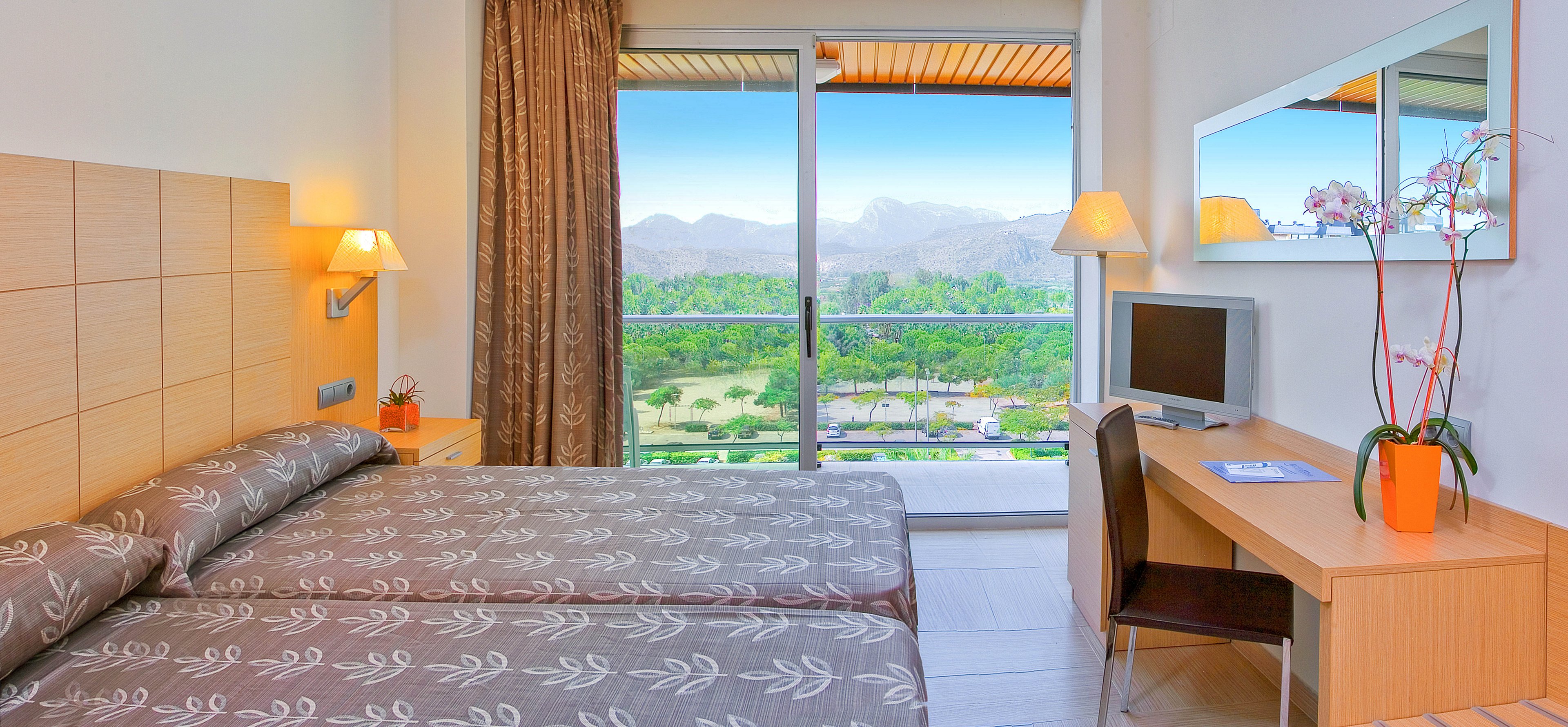 A modern hotel room features twin beds with patterned gray bedding and light wood furnishings. A large glass sliding door opens to a balcony overlooking a lush green landscape and distant mountains under a clear blue sky. A compact desk area with a small flat-screen TV, chair, and orange vase with orchids sits opposite the beds. Warm wall lamps and neutral decor create a clean, inviting atmosphere.