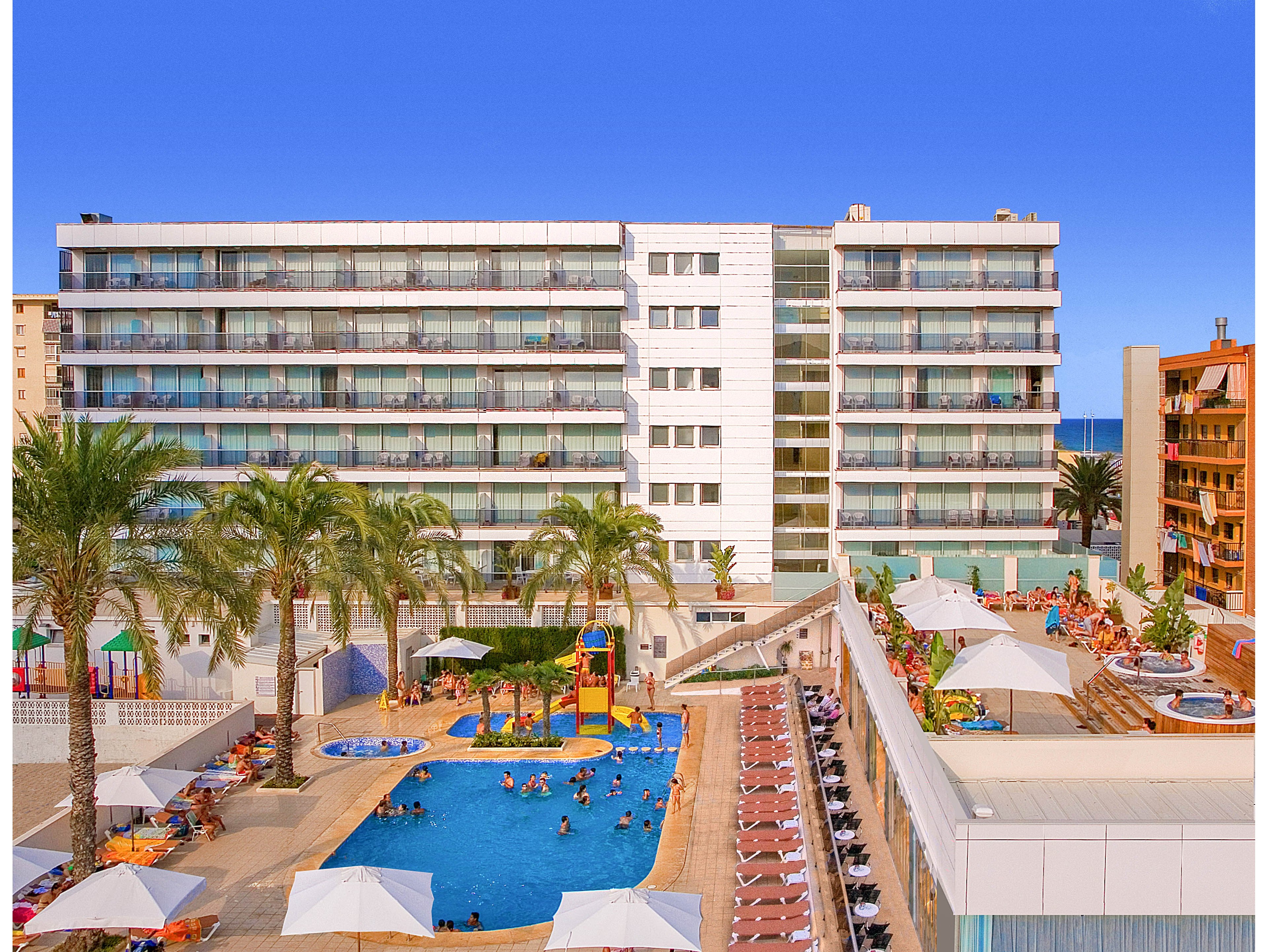 A modern multi-story hotel overlooks a large outdoor swimming pool filled with guests enjoying the water. The scene includes colorful water slides and play structures, surrounded by palm trees and sun loungers. White umbrellas shade rows of deck chairs where people relax around the pool area. In the background, additional buildings and a glimpse of the sea appear under a clear blue sky.