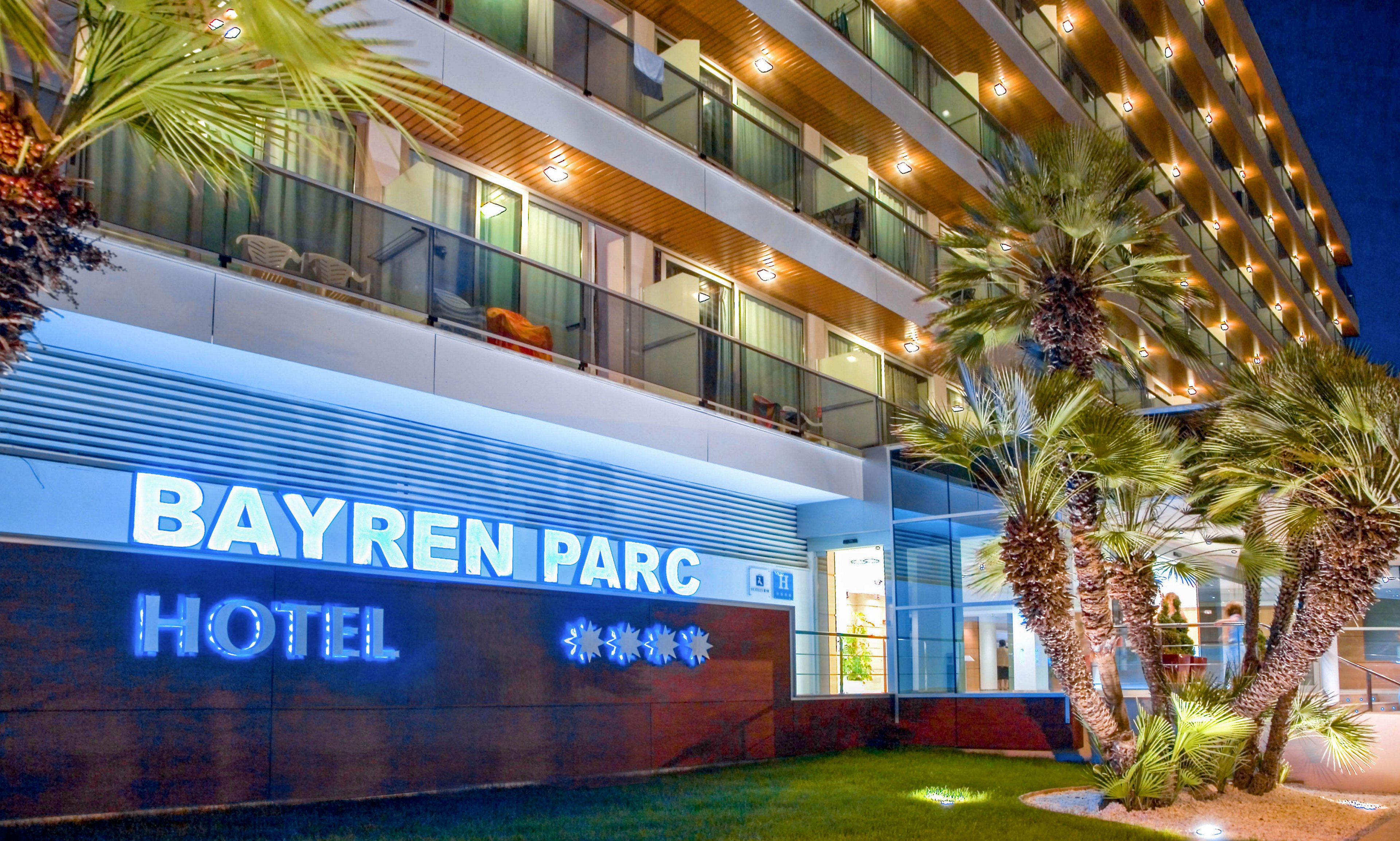 A contemporary multi-story hotel building is illuminated at night with warm balcony lights. The large glowing sign reads "BAYREN PARC HOTEL" with four stars displayed beneath it. Palm trees and landscaped greenery frame the entrance, adding a tropical resort feel. The scene conveys a welcoming, upscale atmosphere suitable for vacation or business stays.