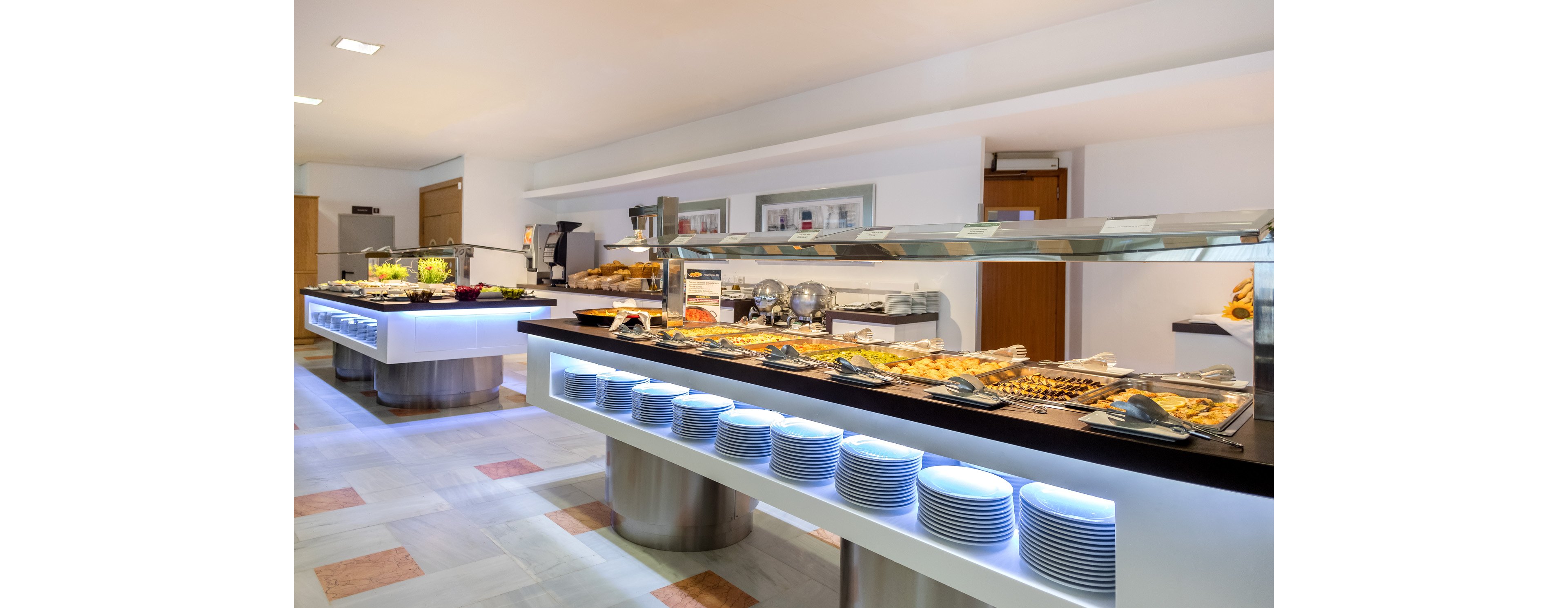 A spacious self-service buffet area features long modern counters filled with assorted hot and cold dishes. Plates are neatly stacked beneath the illuminated serving stations, ready for guests. The bright interior has clean white walls, tiled flooring, and stainless steel accents. Food warmers, salad trays, and dessert options create an inviting dining atmosphere.