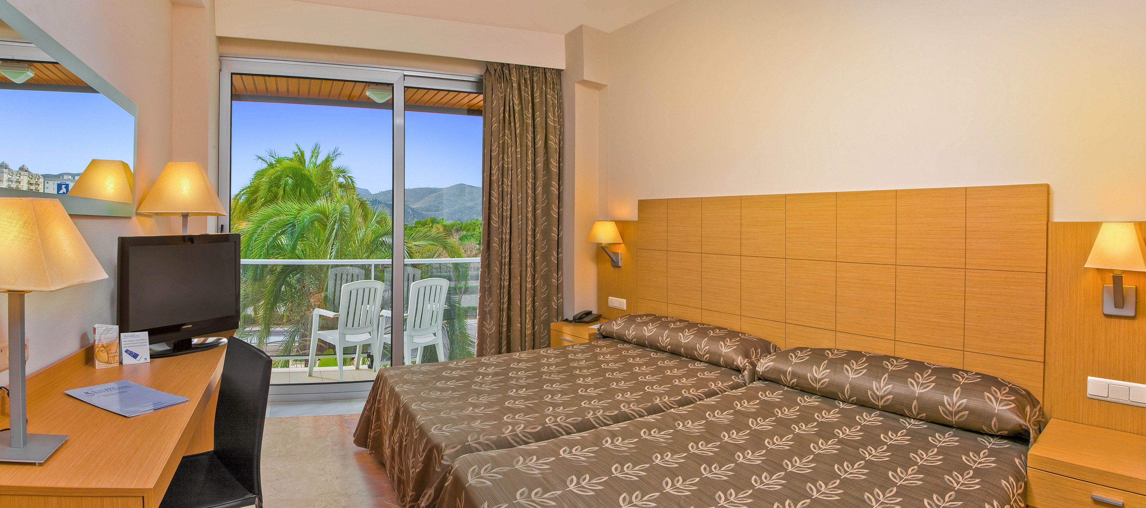 Bright hotel room featuring two single beds with patterned brown bedspreads. A wooden desk with a flat-screen television and lamp sits opposite the beds. Large sliding glass doors open to a balcony with white plastic chairs and a view of palm trees and distant mountains. Warm lighting and neutral tones create a cozy, inviting atmosphere.