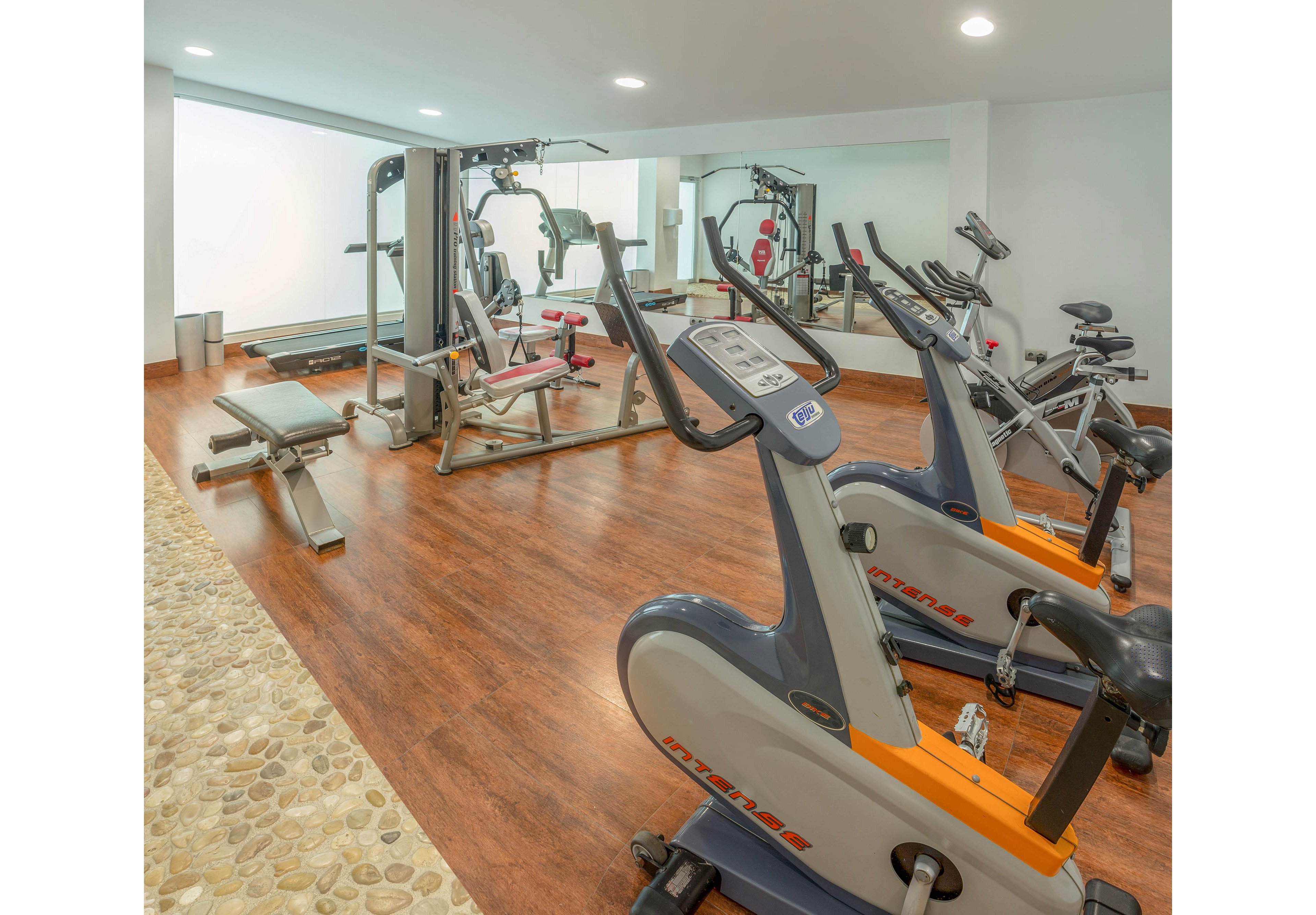 A bright indoor gym features several cardio and strength training machines. Two stationary bikes stand in the foreground facing a mirrored wall. In the background, a multi-station weight machine and bench are arranged on warm wood flooring. Recessed ceiling lights and a white wall create a clean, contemporary fitness environment. A stone-textured walkway borders the workout area, adding a spa-like touch.