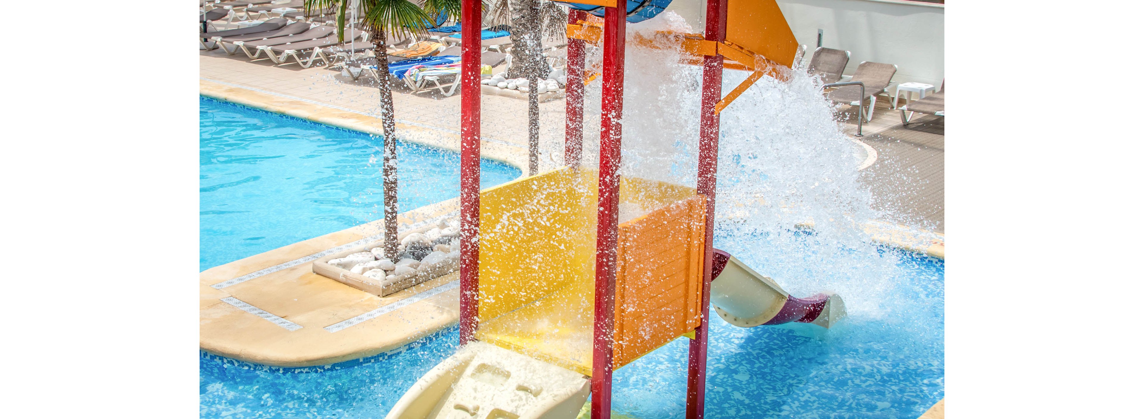 A family relaxes and plays at an outdoor water park pool. Two adults sit at the pool edge while children play under a large tipping bucket that splashes water down. Bright slides, colorful structures, and clear blue water create a fun vacation atmosphere. Lounge chairs and palm trees in the background suggest a resort setting.