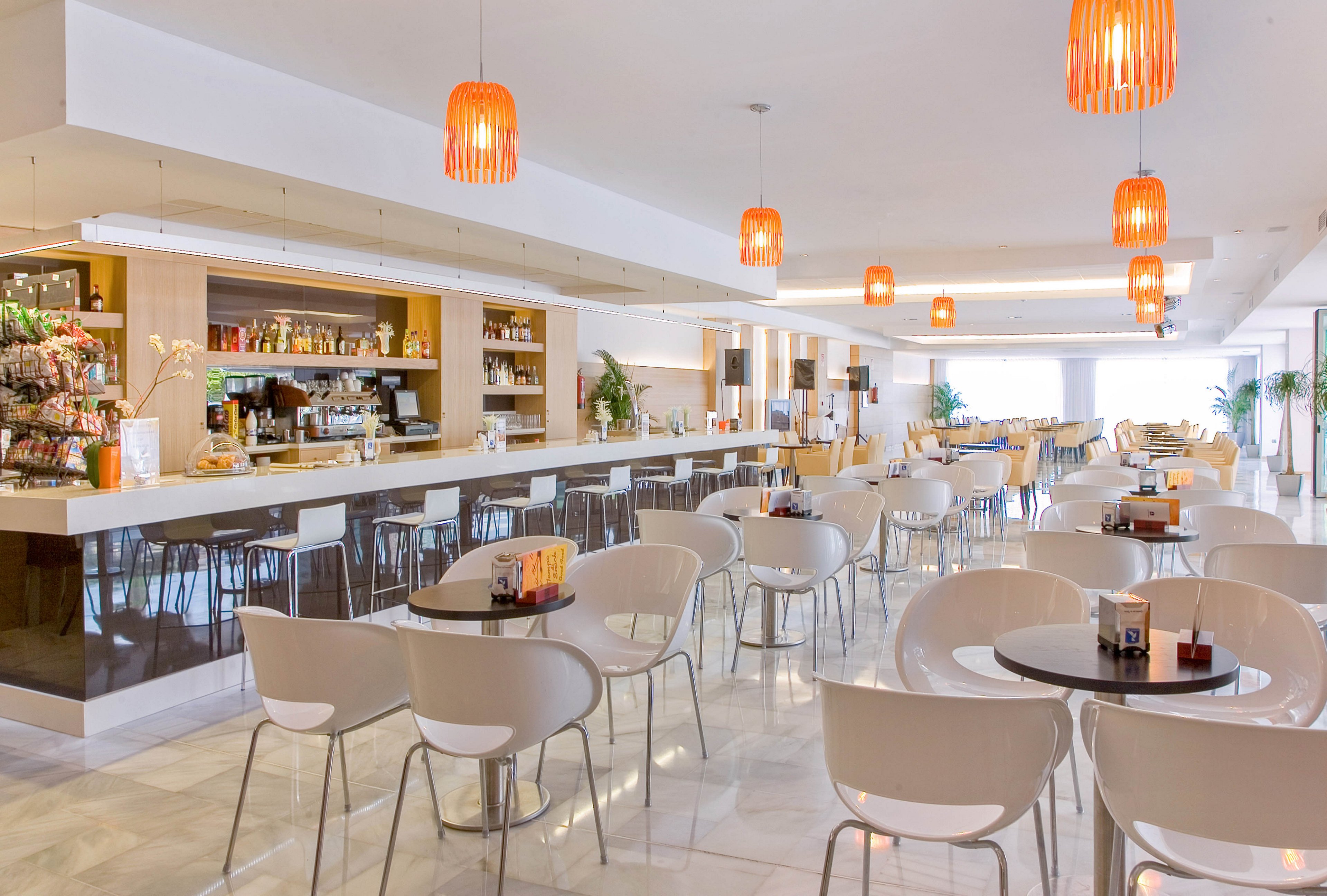 A spacious modern cafe bar features rows of white tables and chairs arranged neatly in front of a long counter. The bar area is stocked with bottles, snacks, and coffee equipment, suggesting a versatile food and drink service. Warm orange pendant lights hang from the ceiling, casting a cozy glow over the otherwise white and neutral interior. Large windows at the back allow natural light to flood the space, enhancing the clean and inviting atmosphere.
