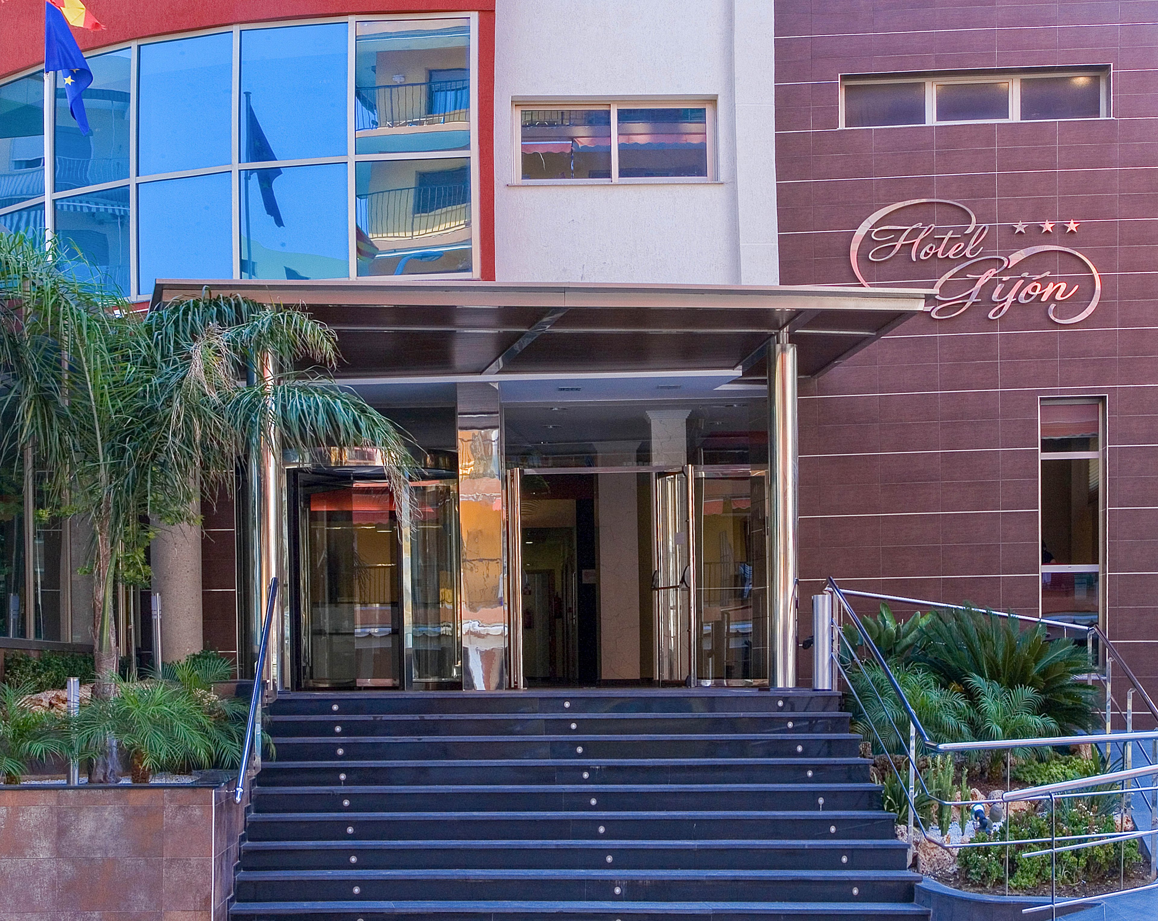 Front entrance of Hotel Gijón with wide dark steps leading up to glass doors. The building facade combines brown panels with white and red sections and reflective blue windows. Palm trees and green plants frame the stairway, adding a touch of nature to the urban setting. A sign reading "Hotel Gijón" with three stars is visible on the wall near the entrance.