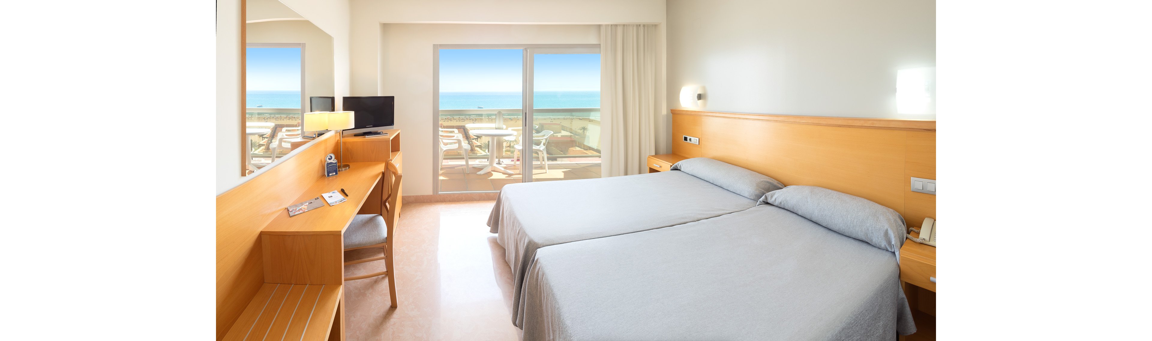 A modern hotel room features two neatly made single beds with light gray bedding. The space includes a wooden desk with a chair, flat-screen television, and simple contemporary furnishings. Large sliding glass doors open onto a balcony with sun loungers and a clear view of the ocean and blue sky. Natural light fills the room, creating a clean, airy, and inviting atmosphere.