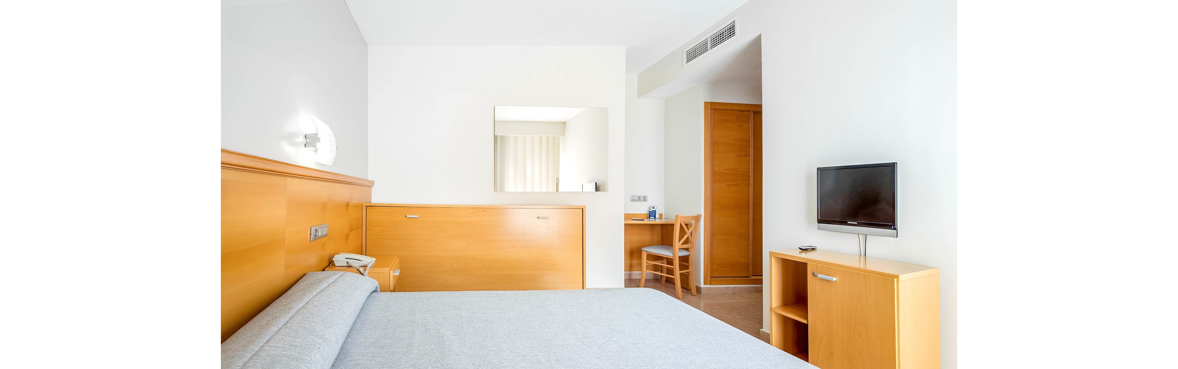 A bright, minimalist hotel room features a neatly made double bed with a light gray bedspread. Light wood furniture, including a headboard, bedside table, and small TV stand, contrasts with the white walls. A flat-screen television sits on the stand, while a wooden chair and desk are positioned near the doorway. Natural light filters in from an unseen window, creating a clean and inviting atmosphere.