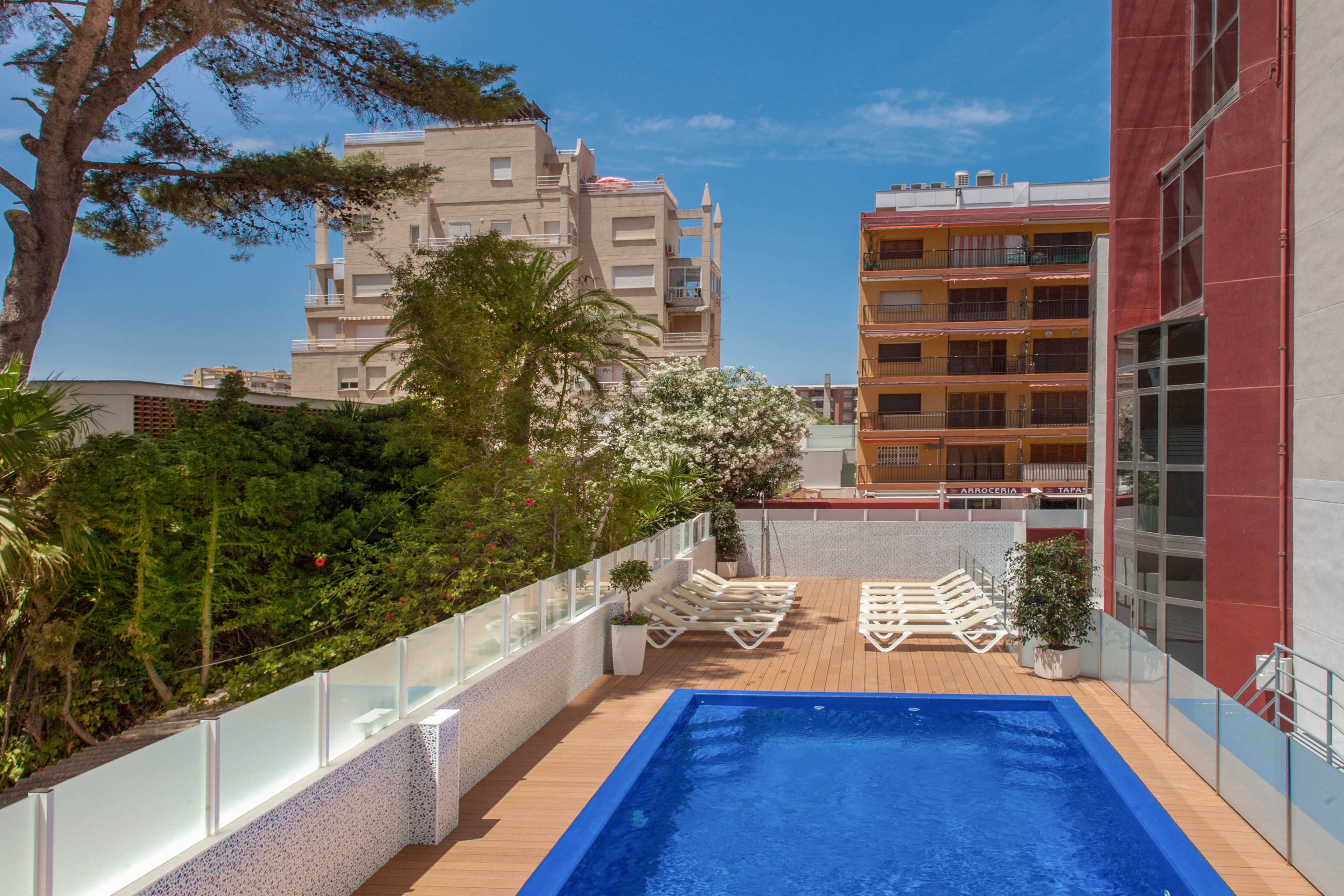 A long rectangular swimming pool is set on an elevated terrace beside a modern building. White lounge chairs line the deck along a glass railing, suggesting a hotel or apartment setting. Tall trees and green hedges border the pool area, with mid-rise urban buildings visible in the background. The bright blue water and clear sky create a sunny, inviting atmosphere.