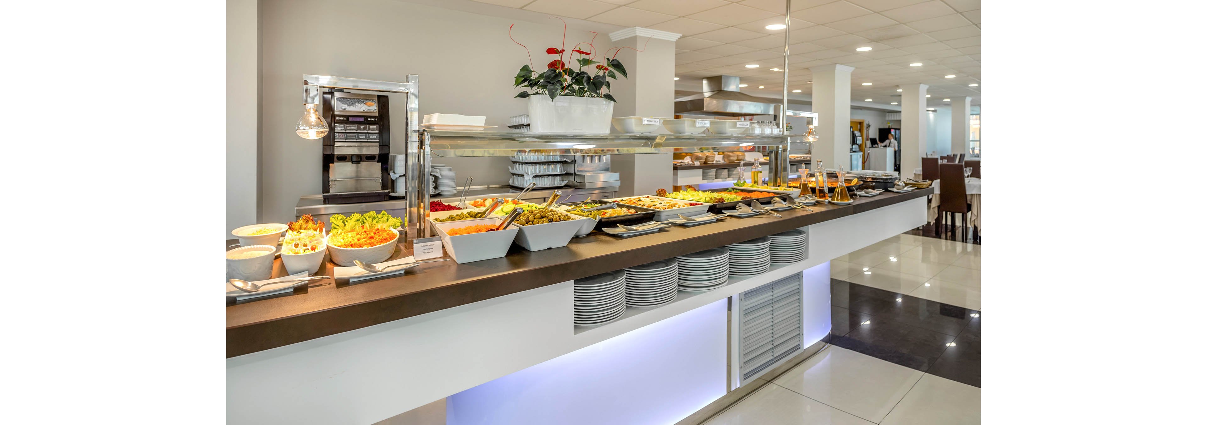 A long buffet counter displays a wide variety of hot and cold dishes in a modern dining area. Fresh salads, fruits, and prepared meals are arranged in white serving trays under warm lights. Stacks of plates are neatly organized beneath the counter, ready for guests. The bright, clean interior features tiled floors, stainless steel equipment, and contemporary decor, suggesting a hotel or restaurant setting.