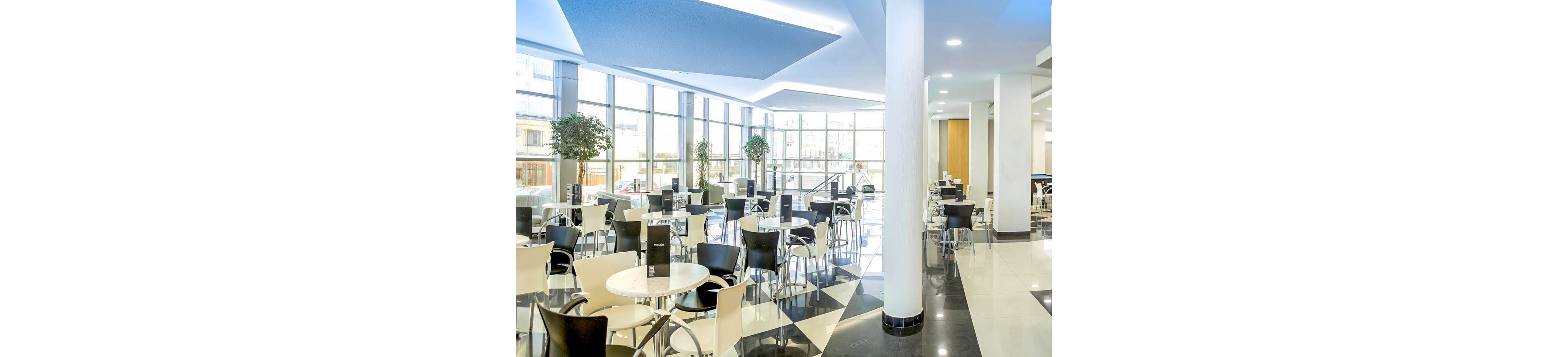 A modern hotel bar features a bright, open lounge with large floor-to-ceiling windows. Two young adults sit on bar stools chatting while a bartender smiles and serves drinks. The interior includes black and white checkered flooring, round tables with metal chairs, and contemporary lighting. Bottles and glasses are neatly arranged behind the bar, creating a clean and inviting atmosphere.