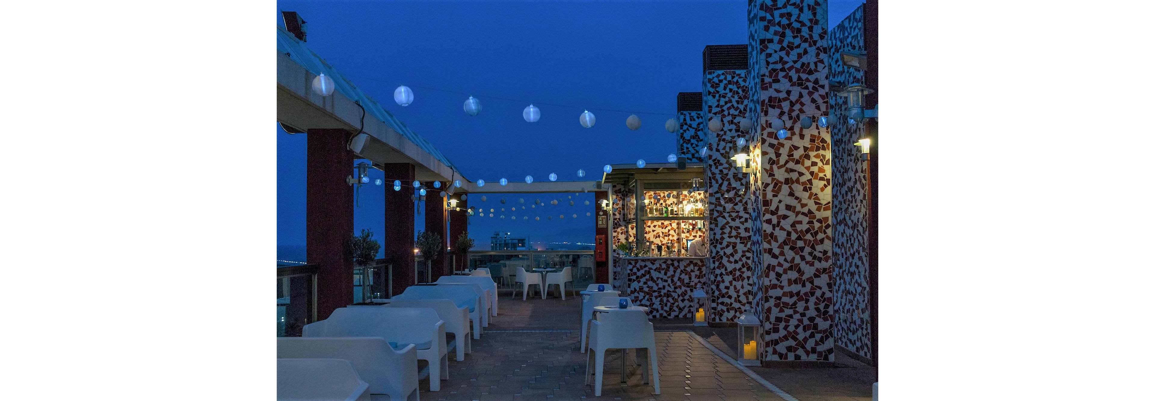A stylish rooftop bar terrace is shown at dusk with modern white chairs and tables arranged along a narrow walkway. String lights and hanging lanterns glow softly above, creating a cozy atmosphere under the deep blue evening sky. A small bar area with illuminated shelves stands near mosaic-covered columns, while candles in glass holders line the floor. In the distance, faint city lights and the horizon are visible beyond the terrace railing.