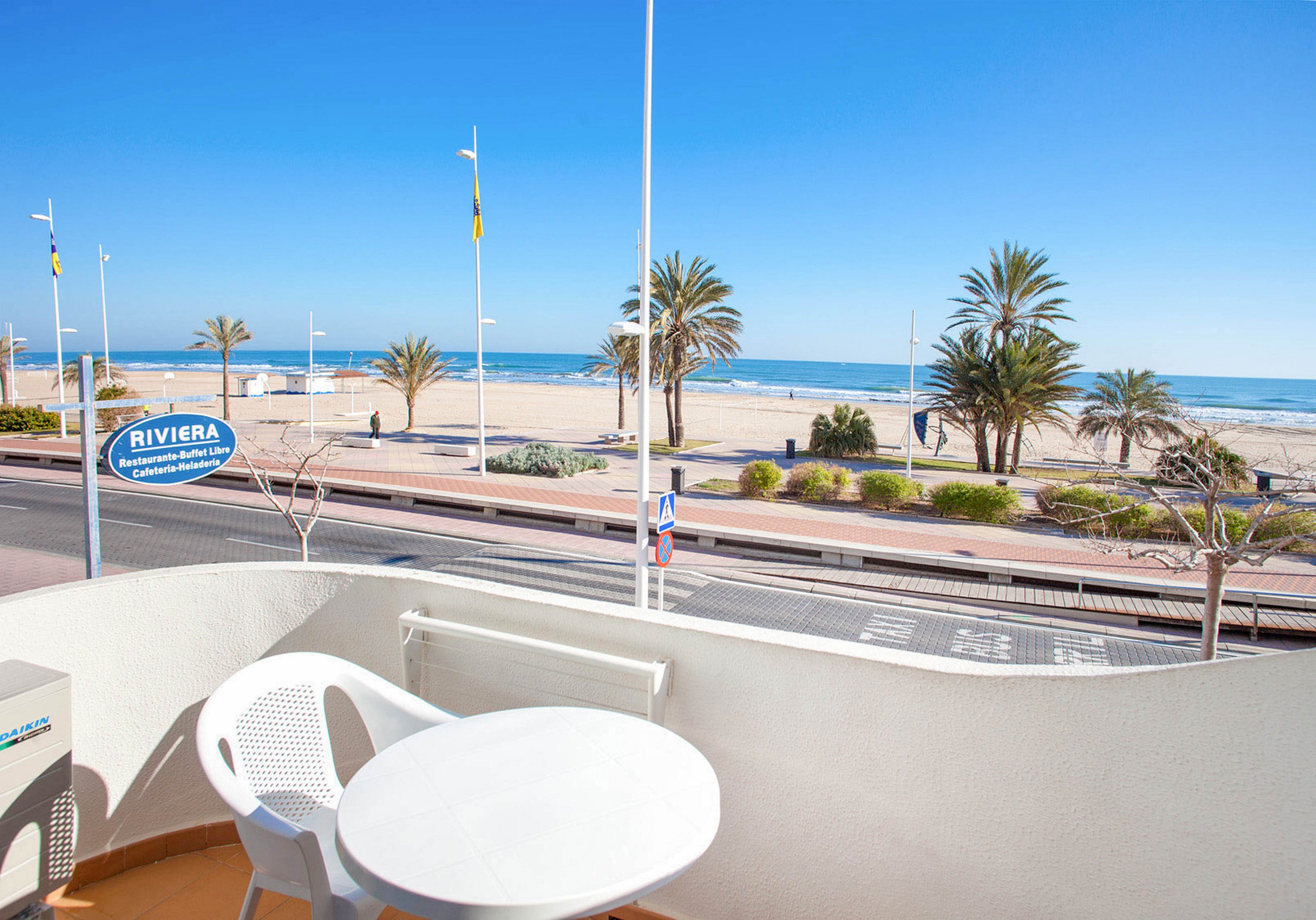 A small white balcony with a round table and plastic chair overlooks a sandy beach and blue sea. The scene includes a coastal promenade with a road, pedestrian walkway, and several palm trees. A bright blue sky and calm water create a clear, sunny atmosphere. A sign reading "Riviera" is visible near the beach, suggesting a seaside establishment.