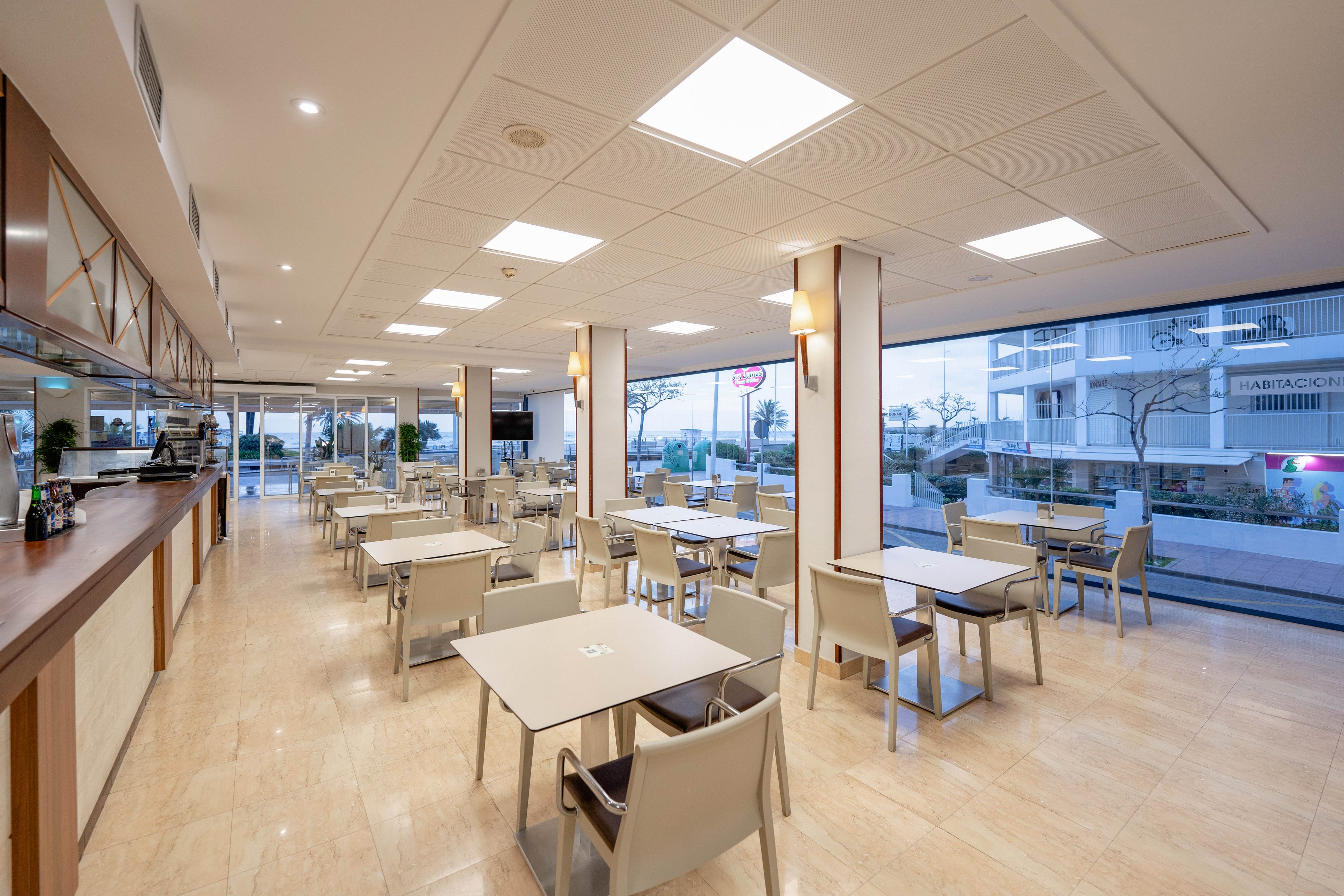 A contemporary restaurant dining area features neatly arranged square tables and beige chairs. The space is empty, highlighting the clean lines and polished light-colored floor. Large floor-to-ceiling windows reveal an urban street and nearby buildings outside. Warm ceiling and wall lights create a bright yet cozy atmosphere, while a long counter runs along one side of the room.