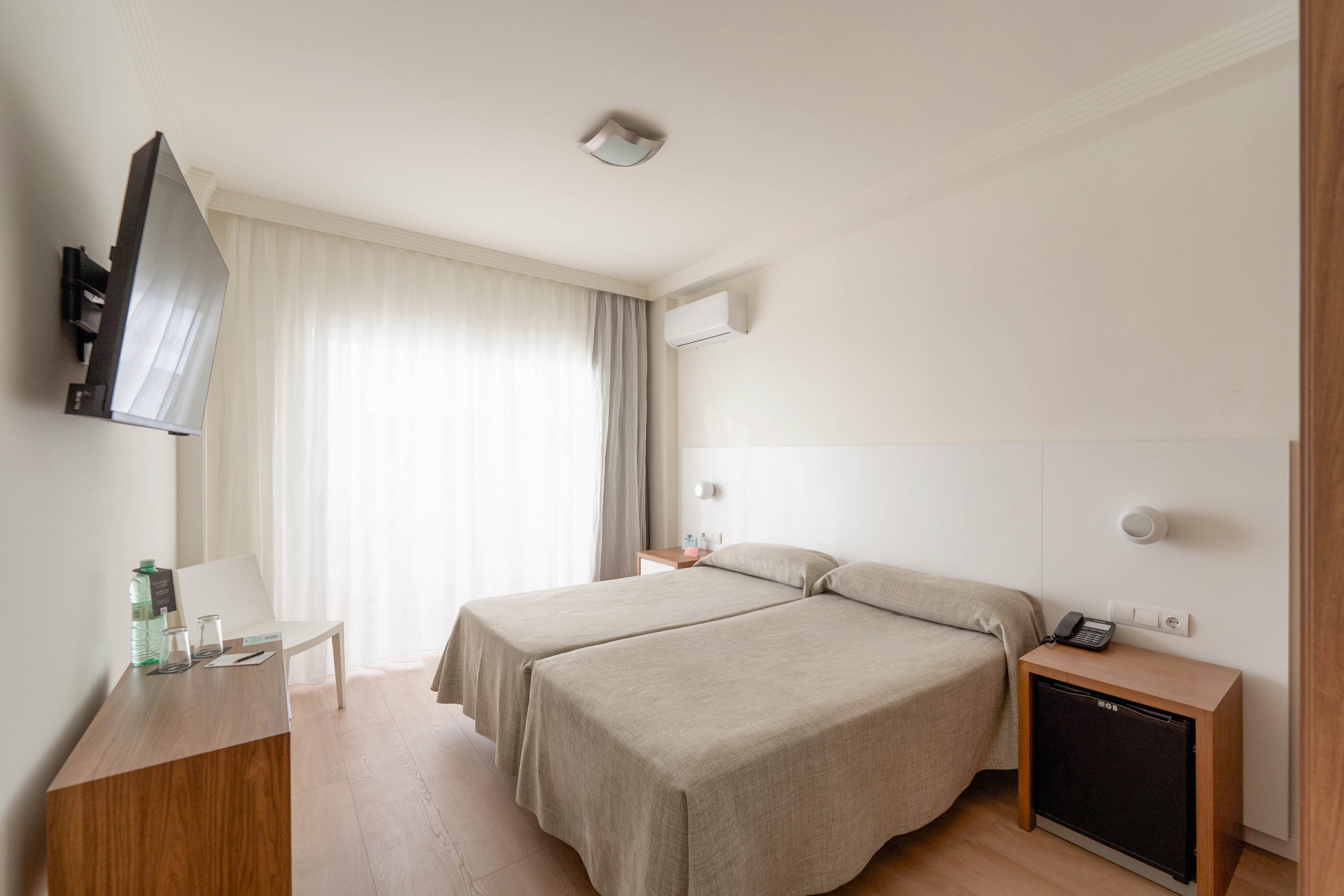 A bright, minimalist hotel room features two neatly made twin beds with beige bedspreads. Soft daylight filters through sheer curtains covering a large window, creating a calm and airy atmosphere. A wall-mounted television faces the beds, and a small wooden table holds water bottles and glasses. A compact refrigerator, telephone, and air conditioning unit emphasize comfort and convenience for guests.