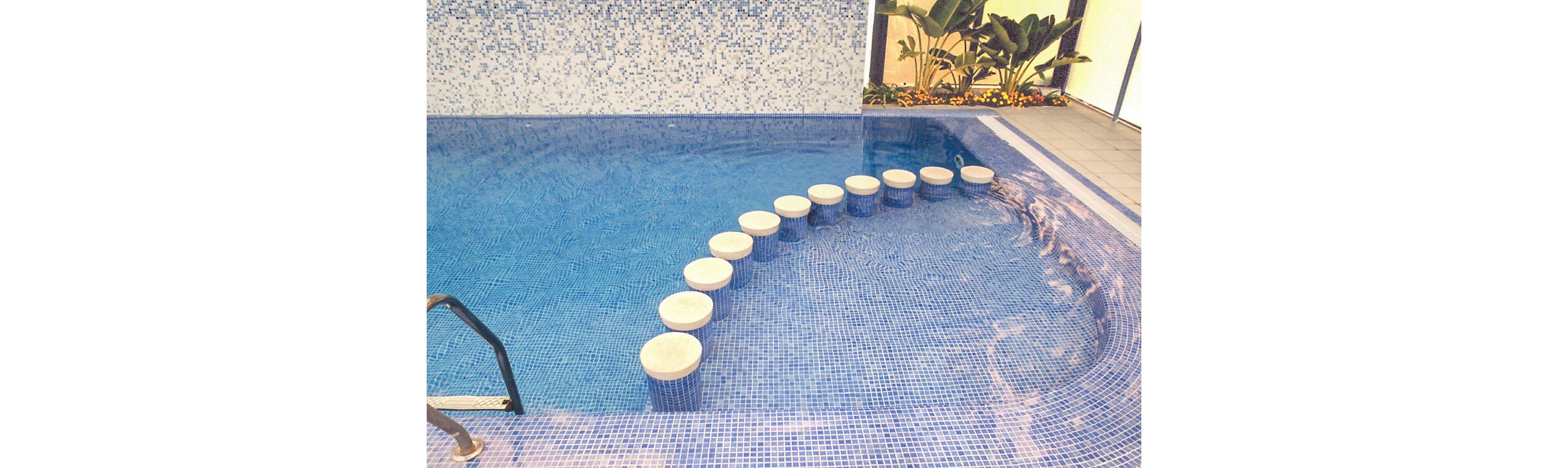A man and woman relax together in a blue-tiled swimming pool. The woman sits on the pool edge while the man leans on a series of round white stepping stools in the water. Mosaic tiles create a gradient pattern from light to dark blue across the pool. In the background, tropical plants and flowers line the poolside area, adding a resort-like atmosphere. The scene feels calm and leisurely, suggesting a vacation or spa setting.