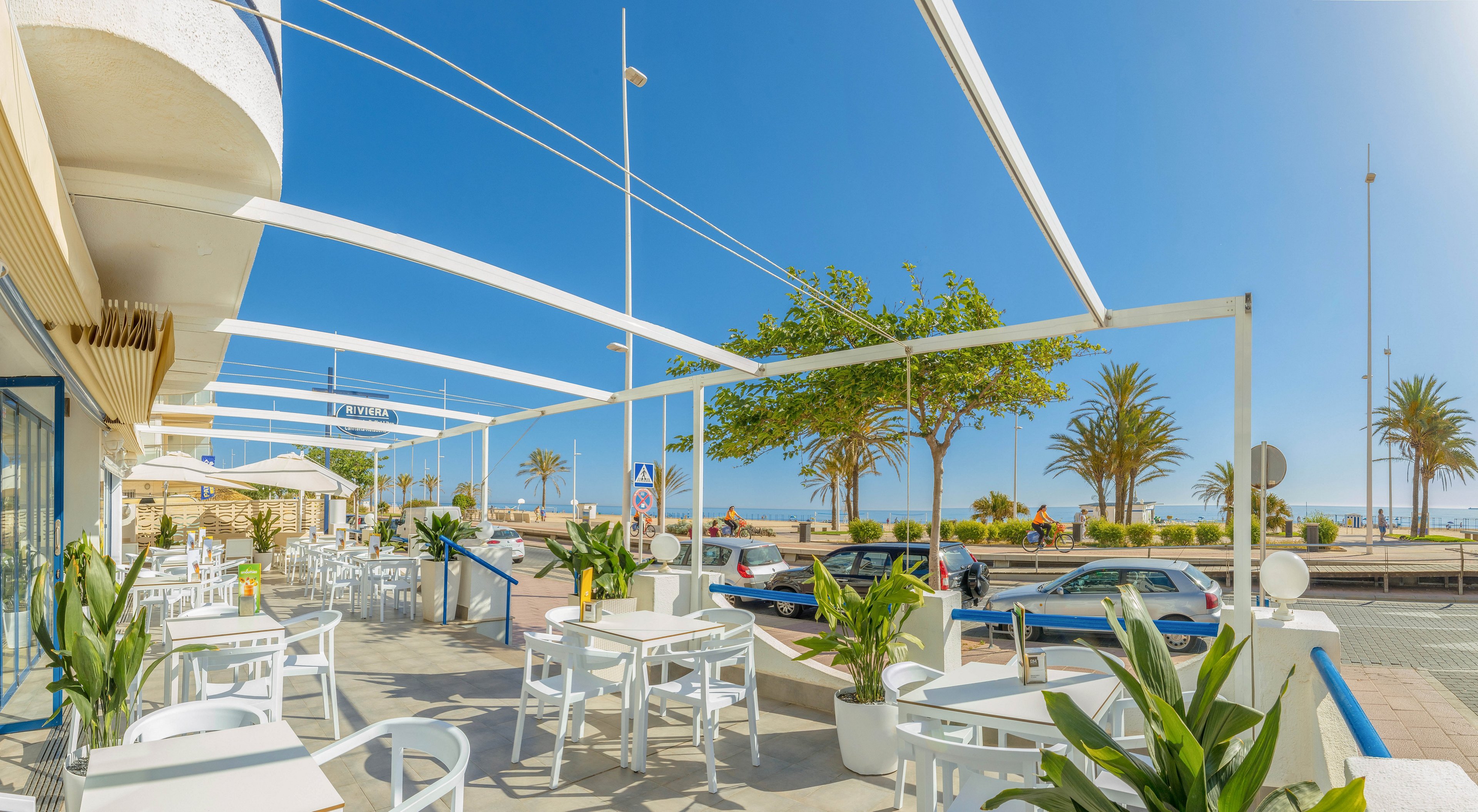 An open-air café terrace with white tables and chairs overlooks a coastal road and sandy beach. Potted green plants decorate the seating area under modern white shade structures. Cars and palm trees line the street, leading toward the bright blue sea and clear sky. The scene suggests a relaxed, warm-weather setting ideal for outdoor dining and leisure.
