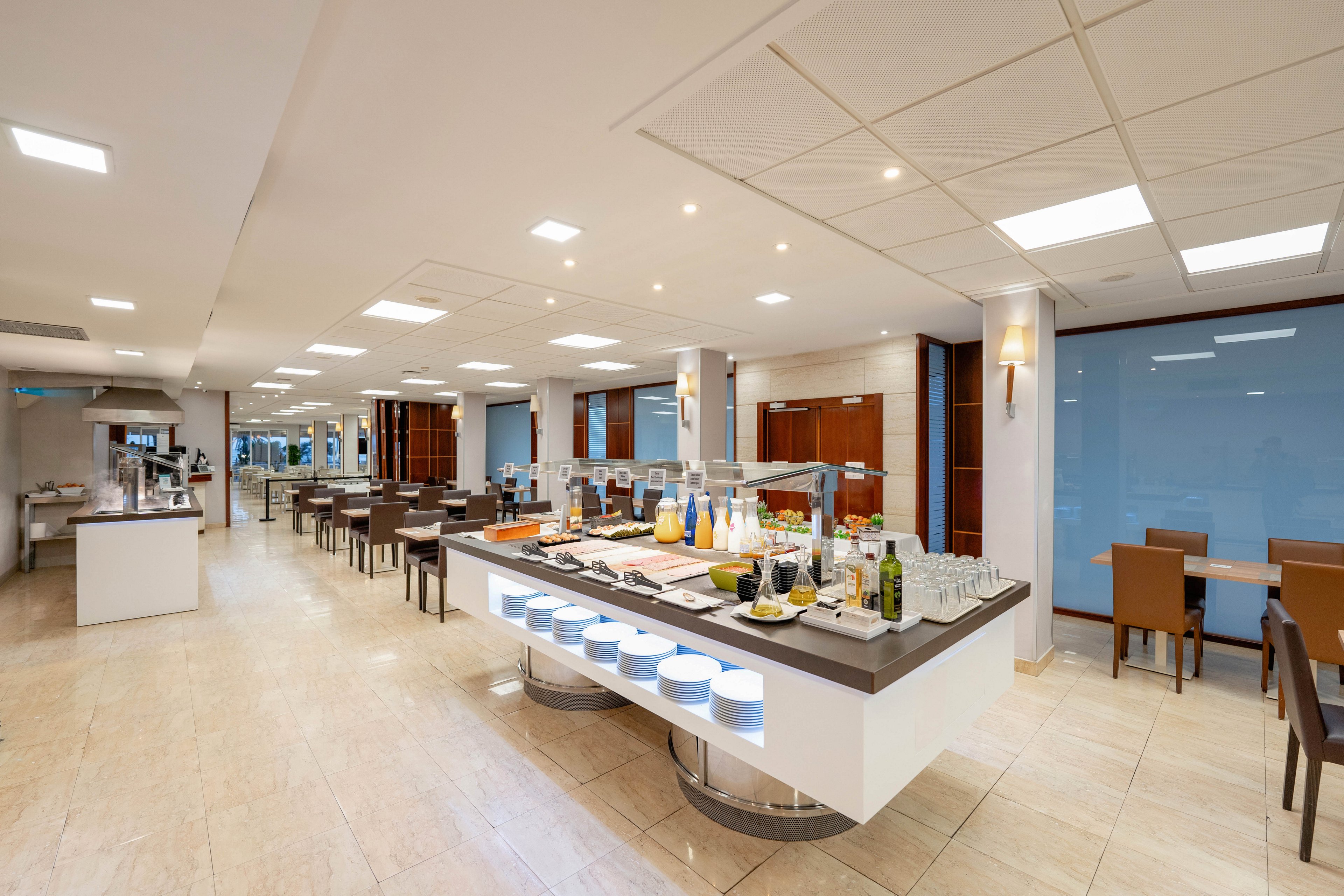 A modern self-service buffet is set up in a bright hotel dining room. The central island displays assorted beverages, fruits, and breakfast items neatly arranged on trays and in glass containers. Rows of tables and chairs extend into the background, creating an organized seating area. Clean lines, neutral tones, and ample lighting give the space a contemporary and inviting atmosphere.