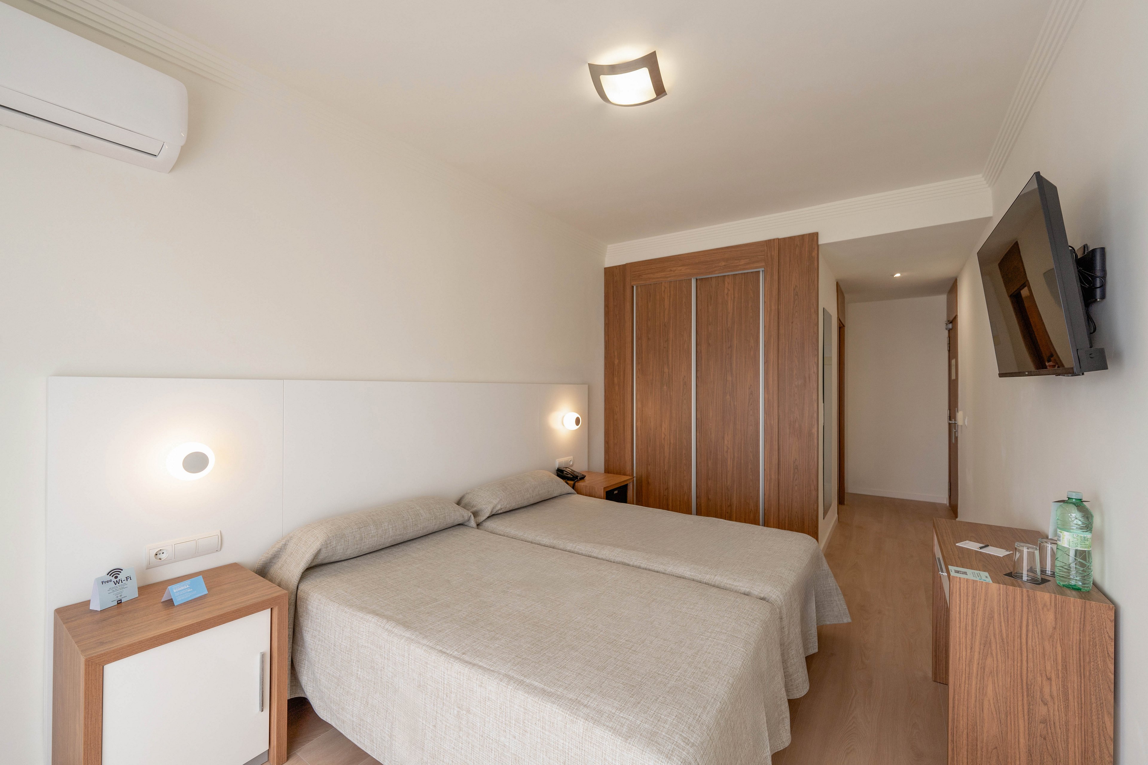 A clean, minimalist hotel room features two neatly made twin beds with light beige bedding. The space includes wooden furniture, such as a bedside table and a wardrobe, complementing the neutral walls and flooring. A wall-mounted television faces the beds, and a split air conditioning unit is installed above. Soft lighting from wall sconces and a ceiling fixture creates a calm, inviting atmosphere.
