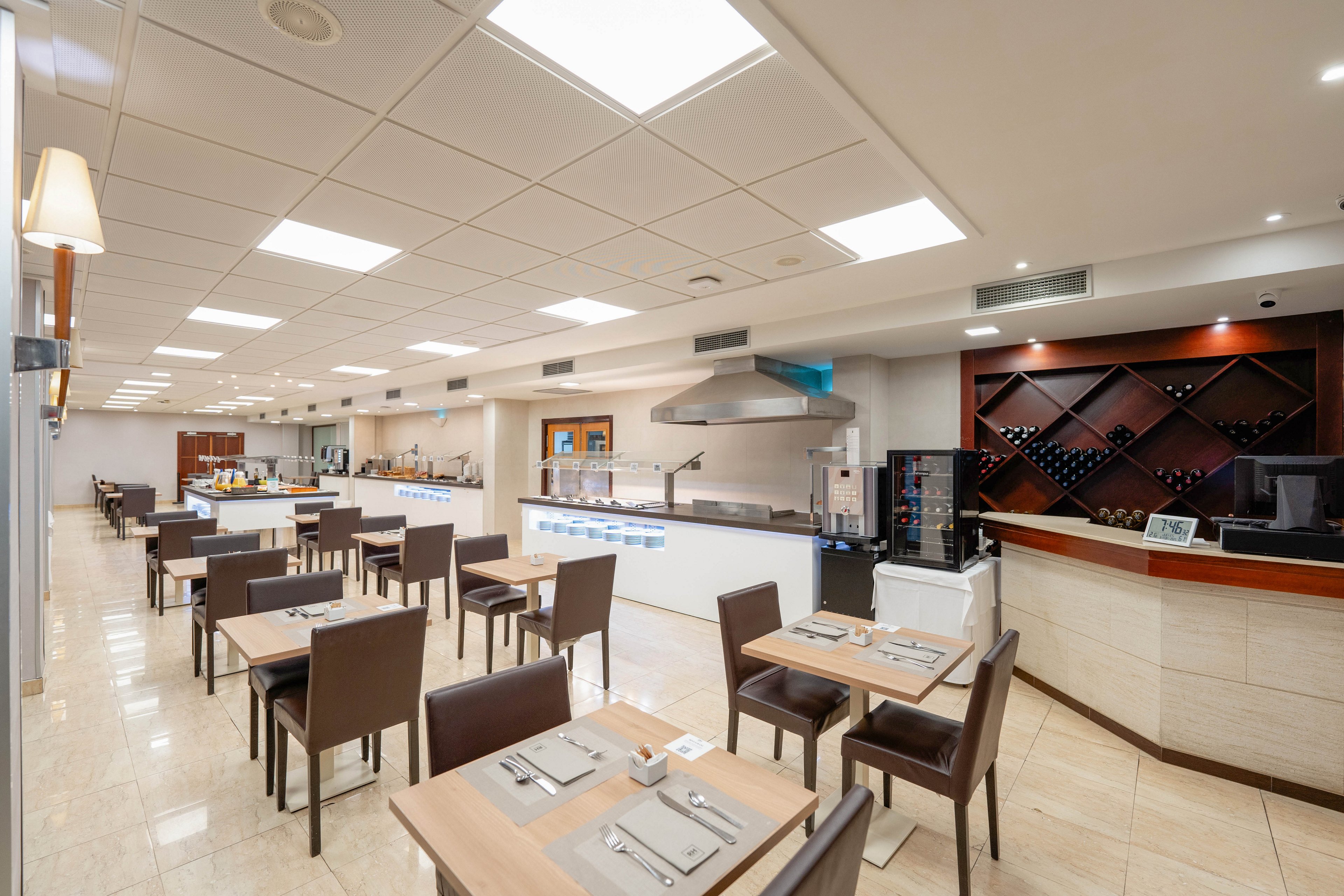 A contemporary restaurant dining area features neatly arranged tables and chairs in a bright, spacious room. The setting includes a long buffet counter with stainless steel equipment and a visible open kitchen area. On the right side, a wine display and service counter add a refined touch to the space. Neutral tones, clean lines, and strong overhead lighting create a minimalist, modern atmosphere.