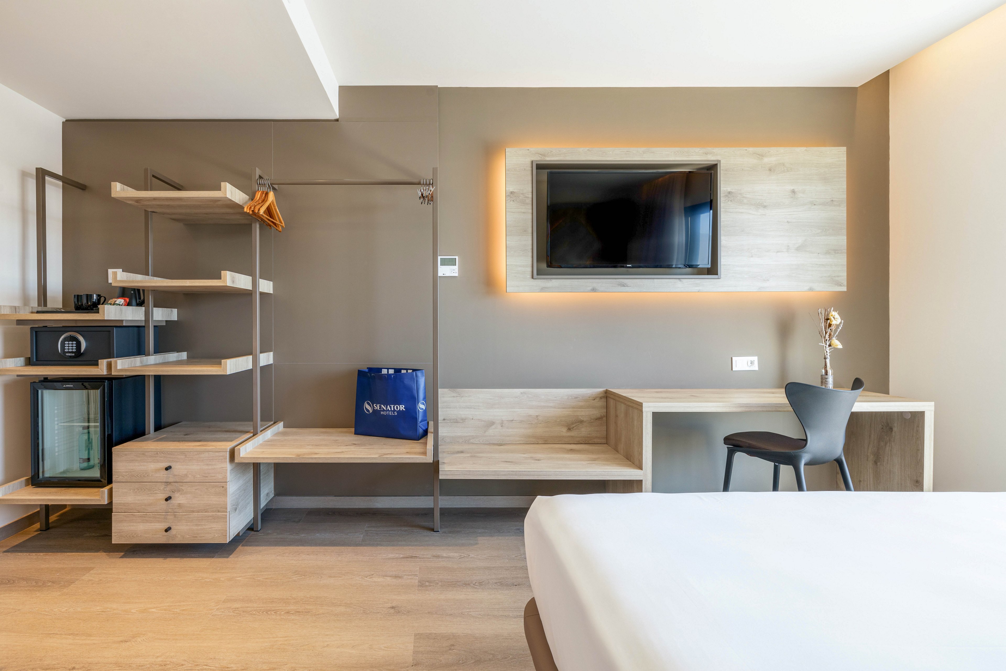 A contemporary hotel room interior features a wall-mounted flat screen television framed by a lighted panel. Below the TV, a built-in wooden bench and desk area with a dark chair provide a minimalist workspace. To the left, open shelving, a small wardrobe rail with hangers, and a compact minibar with a safe create a functional storage area. A neatly made bed is partially visible in the foreground, emphasizing the clean and organized design.
