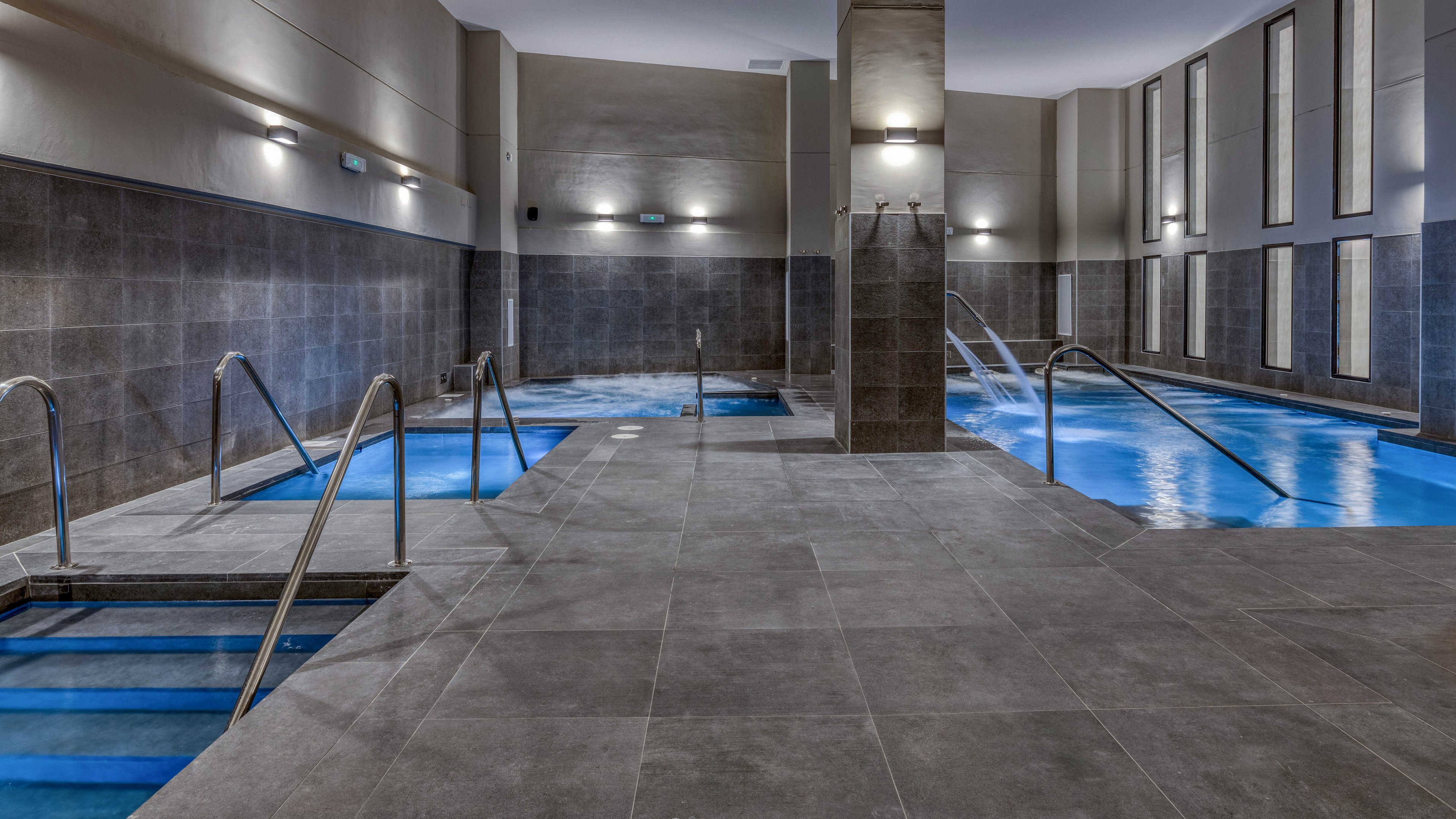 A contemporary indoor spa area features multiple rectangular pools surrounded by sleek gray tiles. Stainless steel handrails lead into the illuminated blue water, creating a clean and inviting atmosphere. Tall vertical windows and wall sconces provide soft, diffused lighting throughout the space. The overall design feels calm, minimalist, and luxurious, ideal for relaxation and wellness.