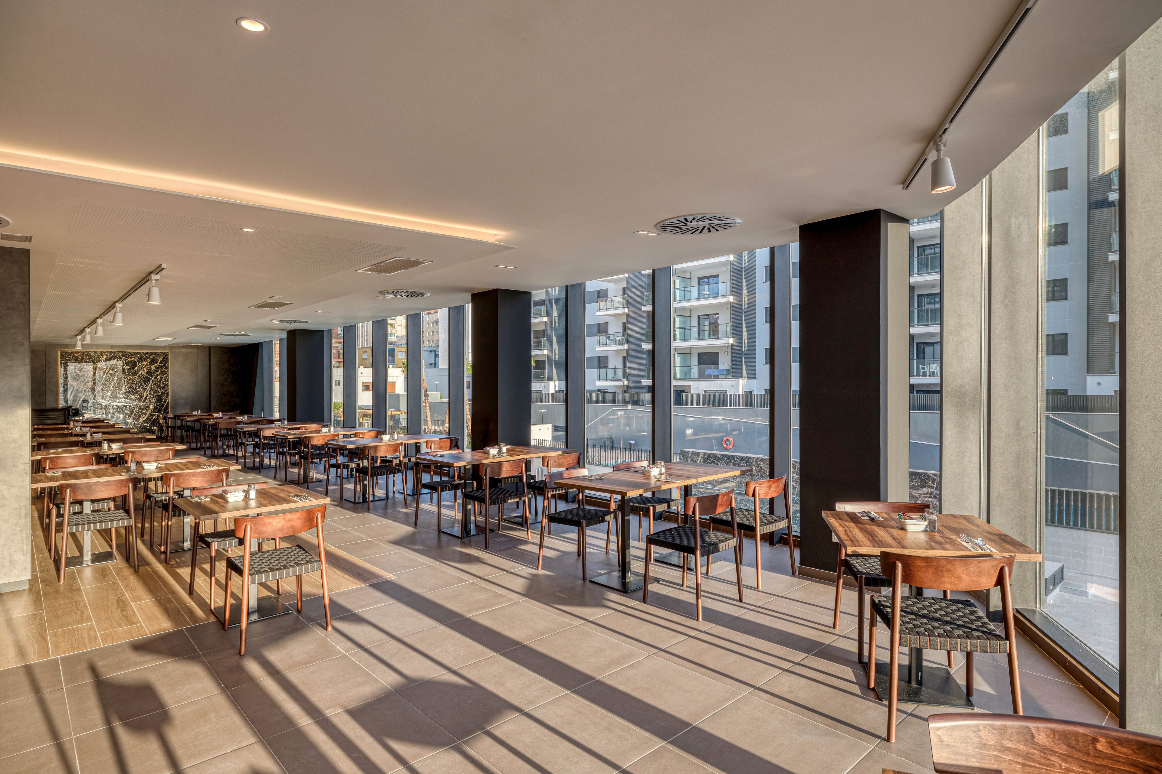 A contemporary restaurant dining room features rows of wooden tables and chairs neatly arranged in an open layout. Floor-to-ceiling windows line one side of the space, revealing a cityscape of modern apartment buildings outside. Warm sunlight streams through the glass, casting long shadows across the tiled floor and highlighting the clean, minimalist design. The room appears quiet and empty, ready to welcome guests for a meal service.