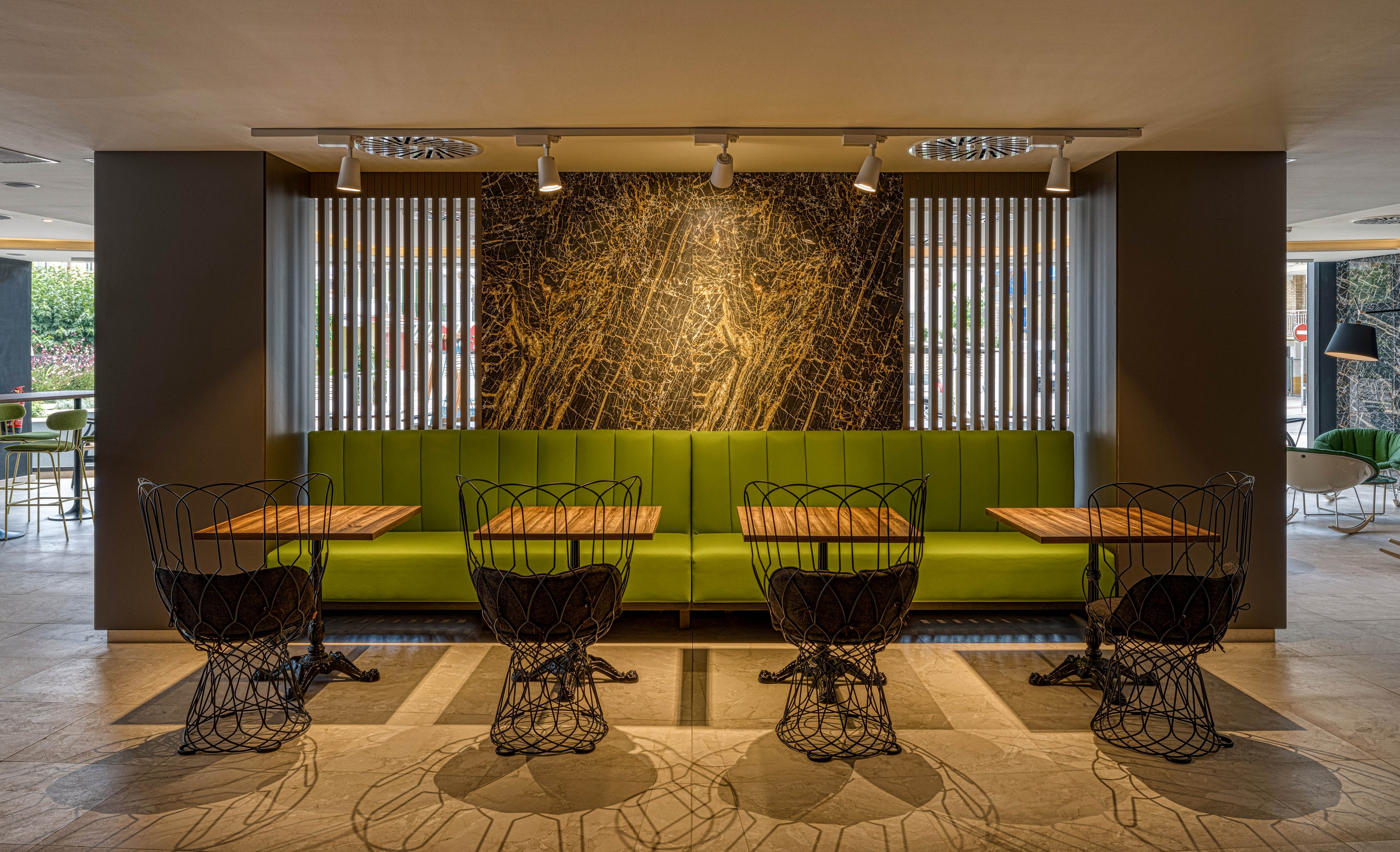 A contemporary cafe interior features a long green banquette framed by dark walls and a textured stone accent panel. Four small wooden tables are paired with sculptural black wire chairs arranged symmetrically. Vertical wooden slats and recessed ceiling lights create a warm, inviting ambiance. Additional seating and natural light are visible in the background, suggesting an open, social space.